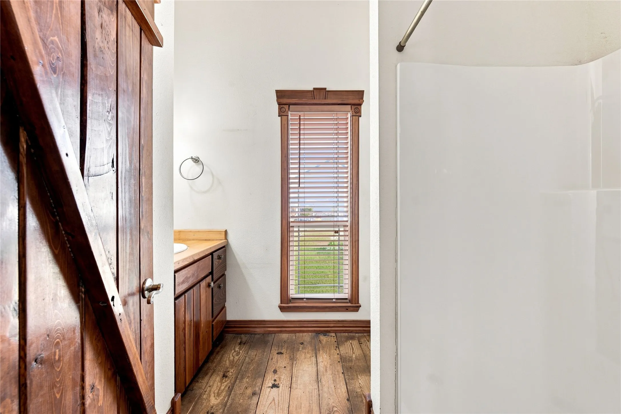 Wood-finish flooring, wood vanity with integrated sink, wood window trim, and white shower surround