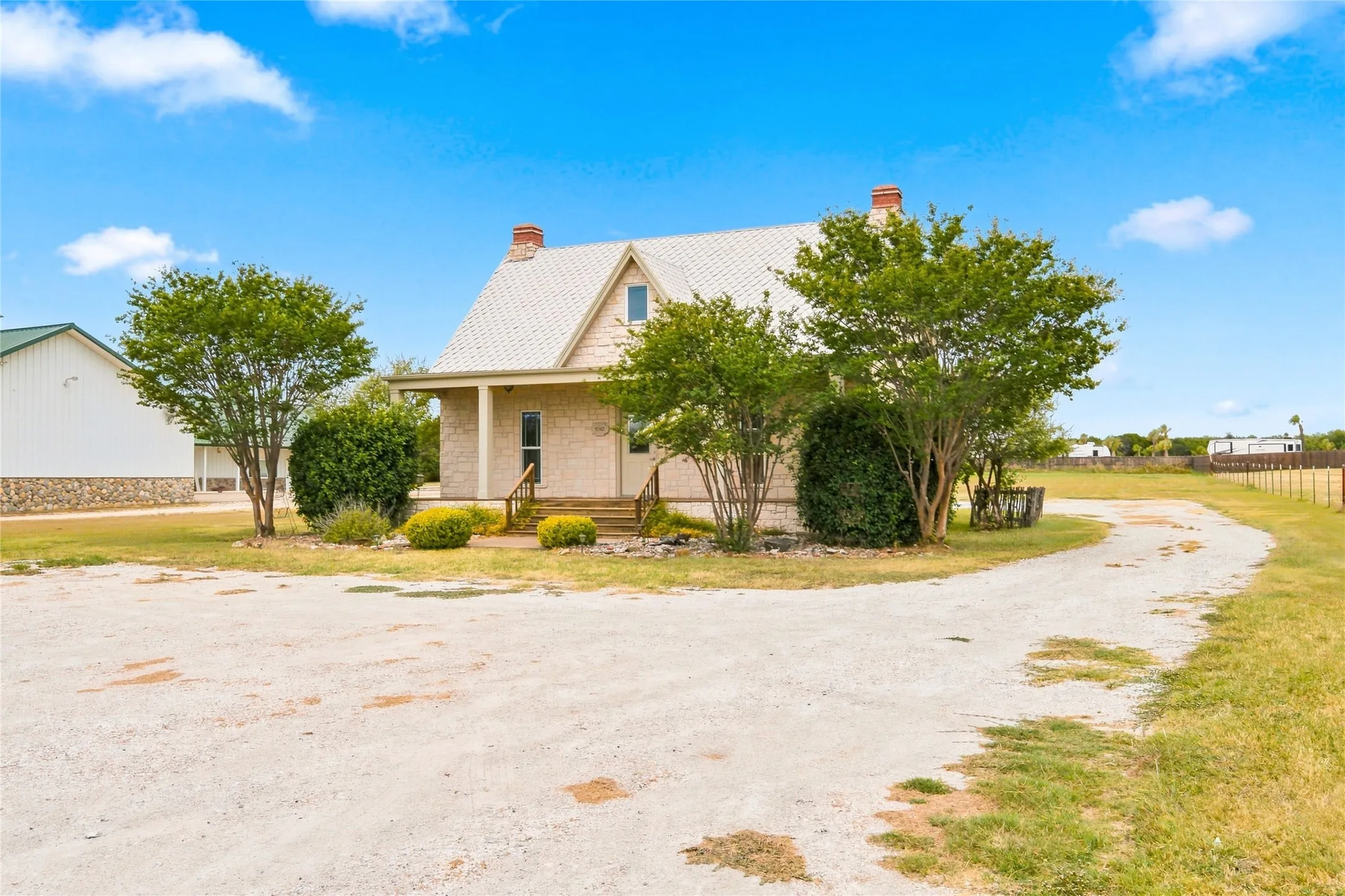 Gable roofline with a metal roof, front porch with columns, mature landscaping, and a gravel driveway