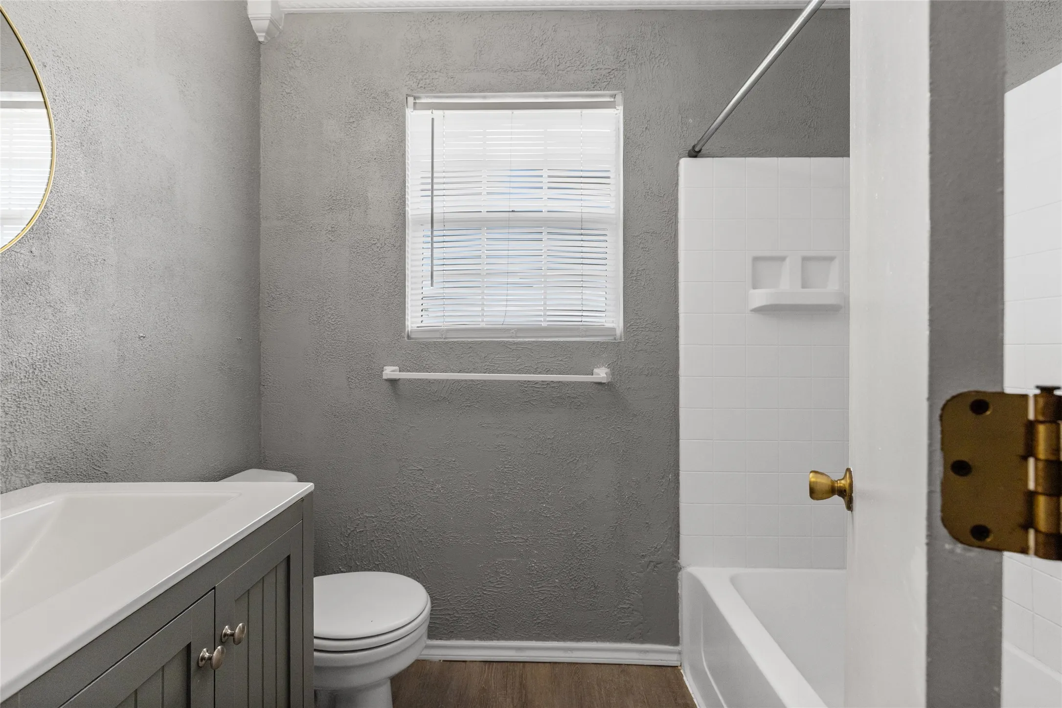 Compact bathroom featuring a white porcelain tub, a window with blinds, and wood-finish flooring