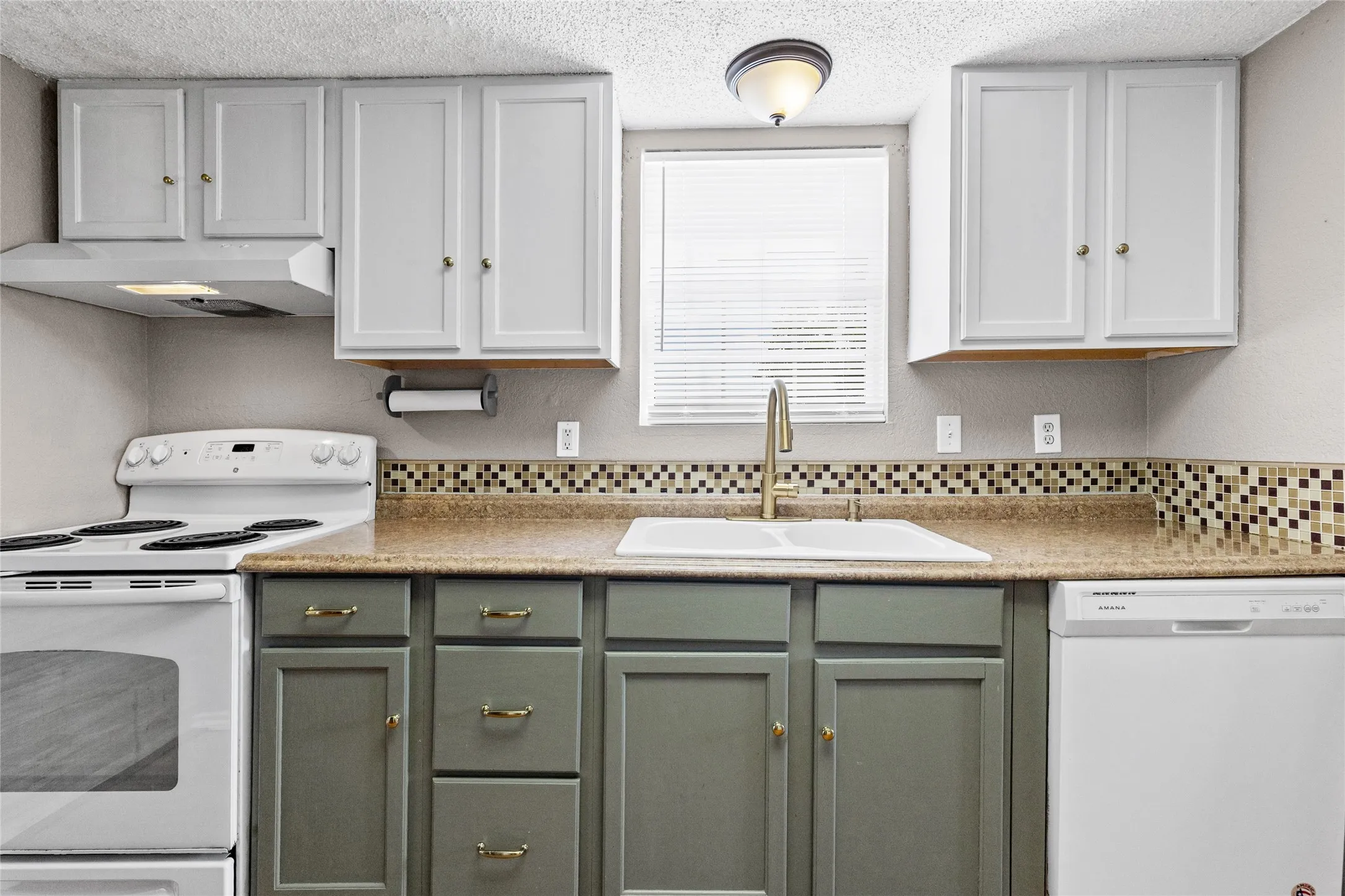 Two-tone cabinetry with upper white cabinets and lower gray cabinets, mosaic tile backsplash, countertop with integrated sink, modern gold-tone faucet, and white appliances