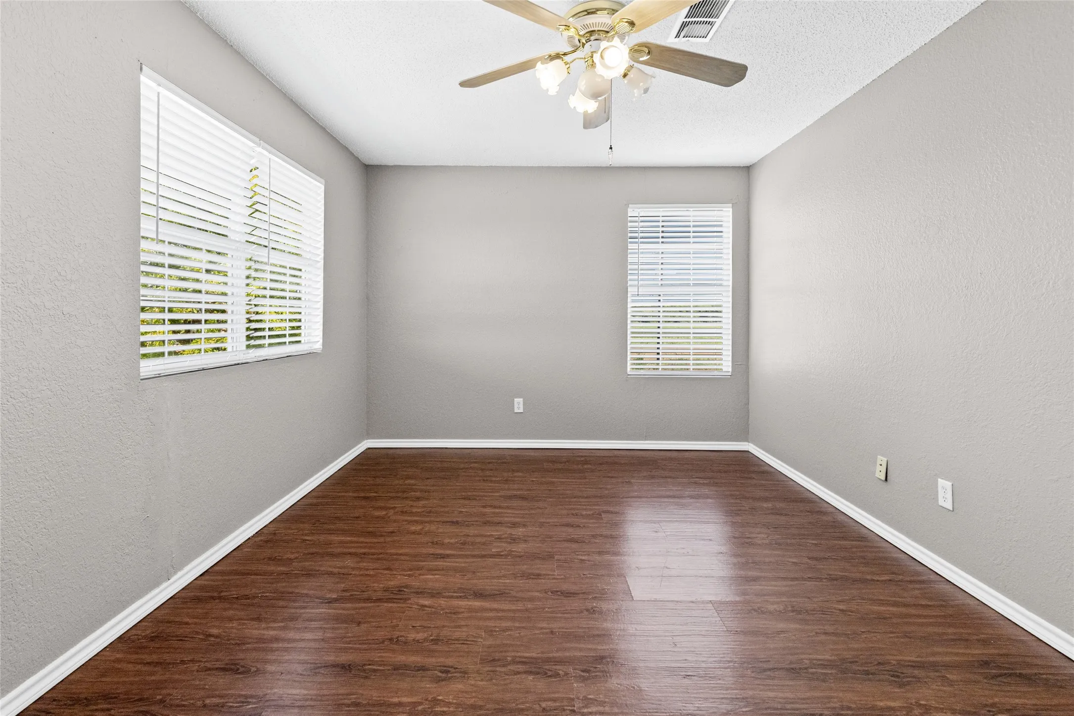 Wood-finish flooring with white trim, light gray walls, multiple windows with blinds, and a ceiling fan with integrated lighting