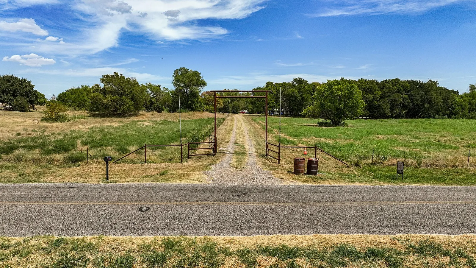 Gravel driveway with a metal entry gate, bordered by open fields and mature trees