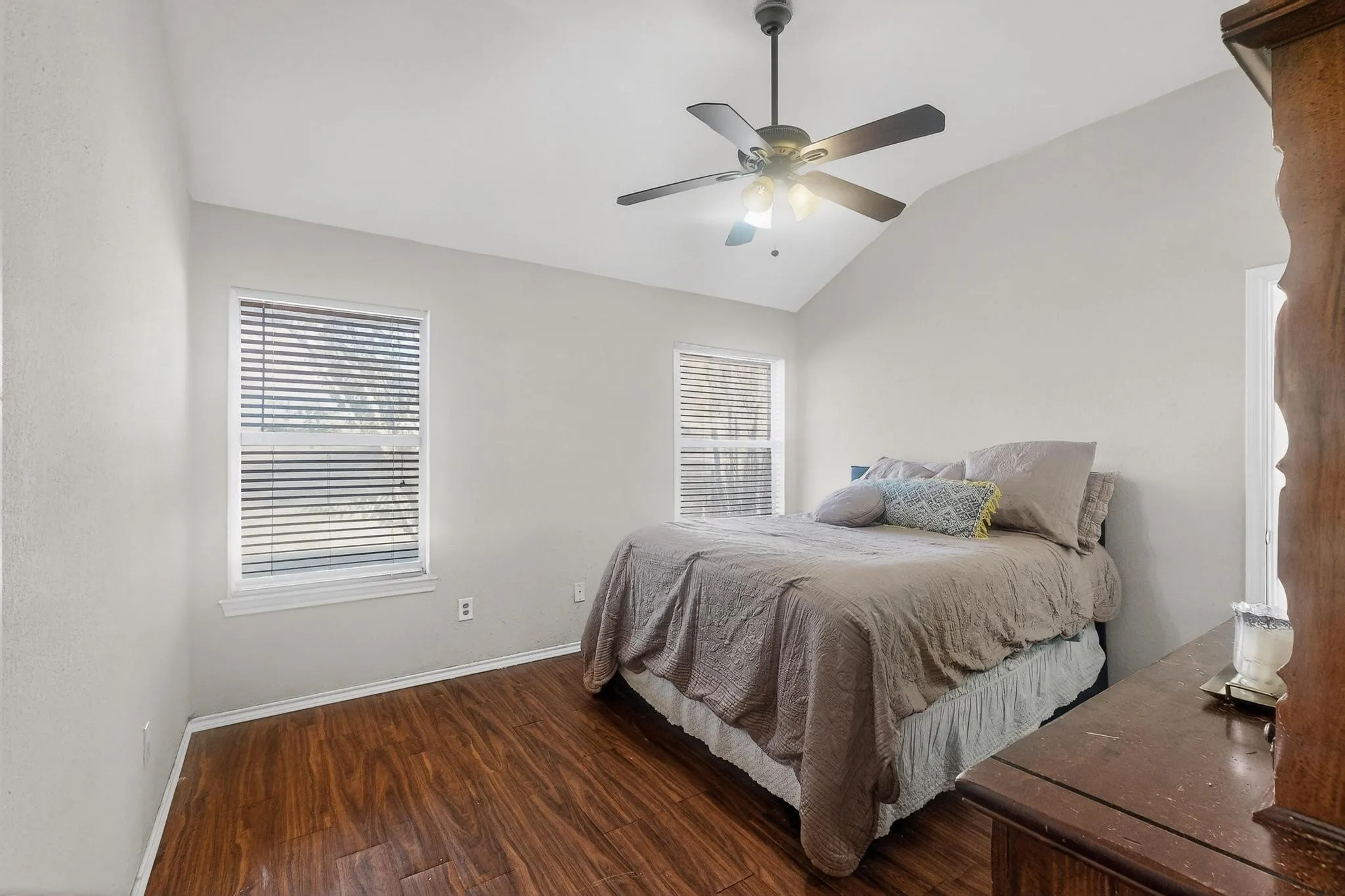 Vaulted ceiling, wood-finish flooring, ceiling fan, two windows with blinds, and neutral wall paint