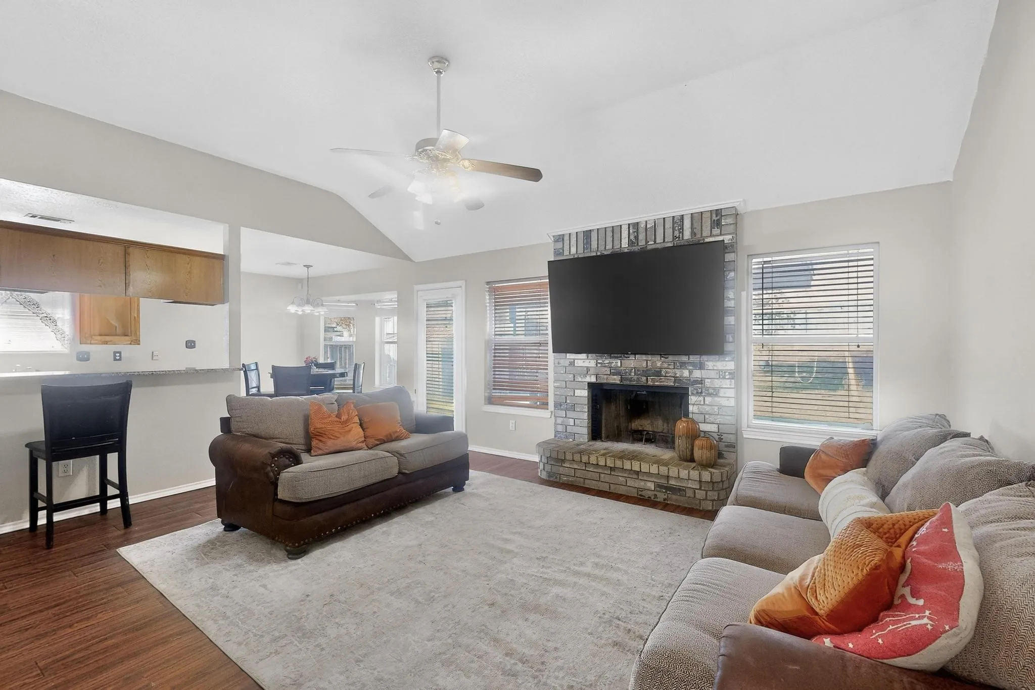 Vaulted ceiling with a ceiling fan, brick-clad fireplace, wood-finish flooring, multiple windows with blinds, and a breakfast bar