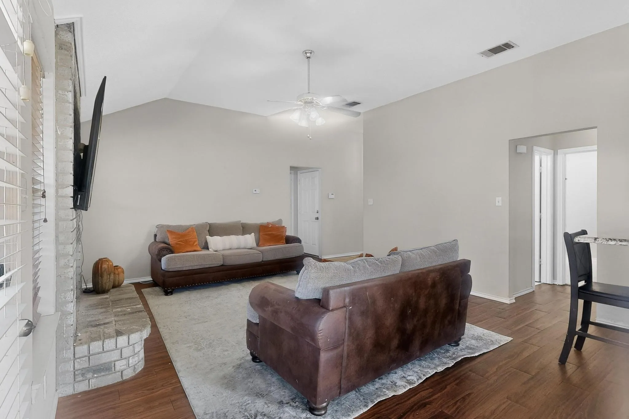 Vaulted ceiling, wood-finish flooring, brick fireplace, ceiling fan, and light gray wall paint