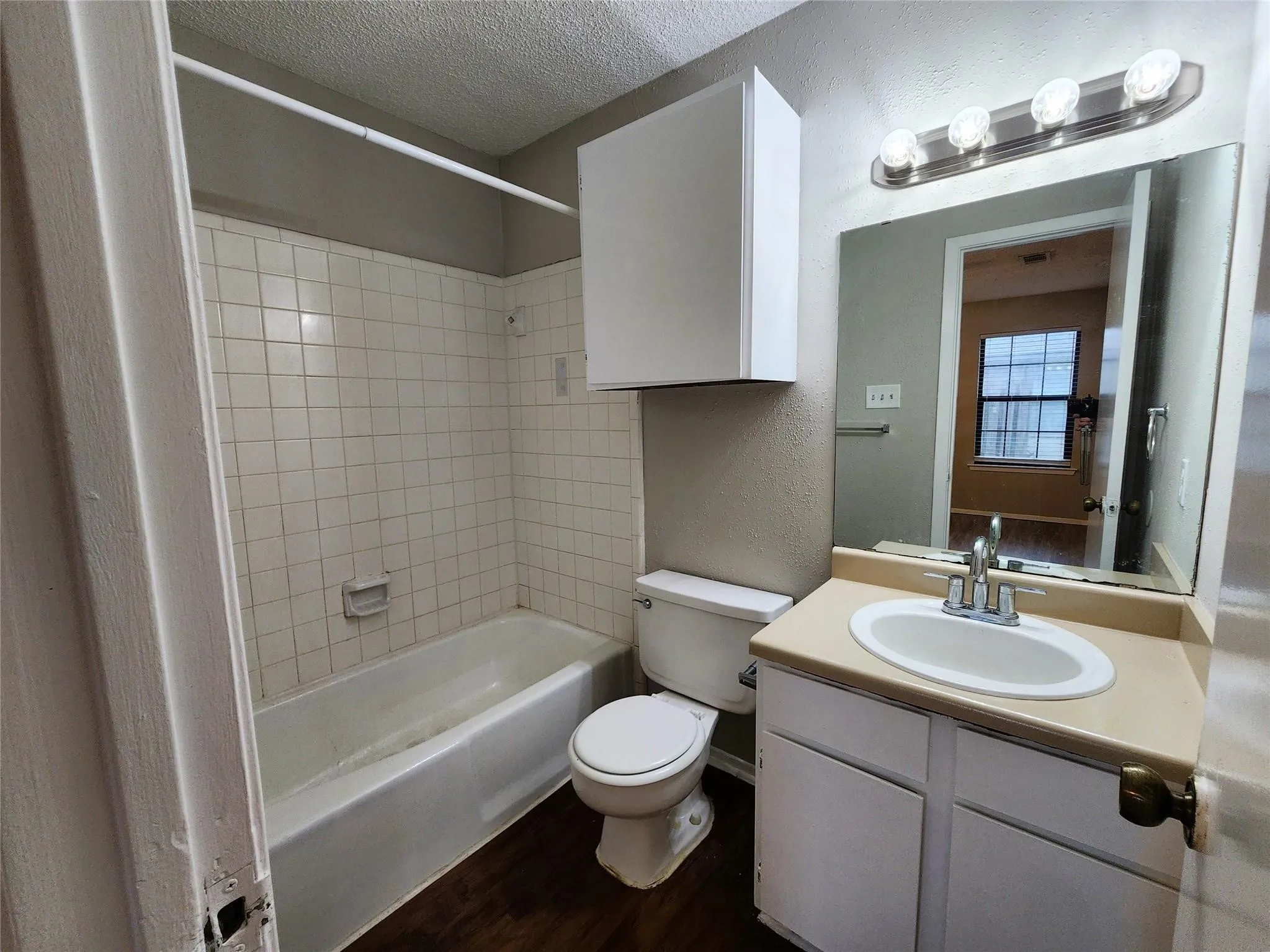 Bathroom featuring a white vanity with an integrated sink, a vanity mirror with overhead lighting, a white toilet, a bathtub with tile surround, and wood-finish flooring