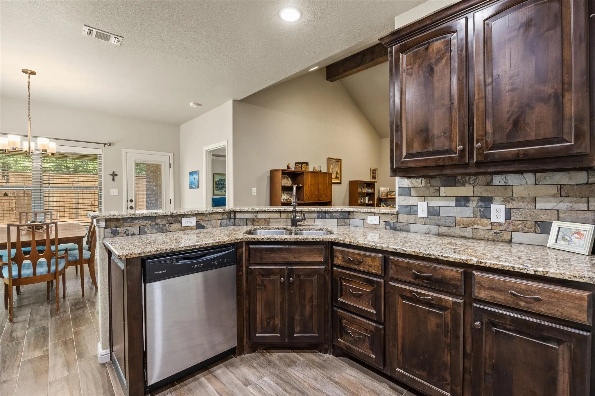 Kitchen featuring wood-look tile flooring, dark wood cabinetry, granite countertops, a stainless steel dishwasher, and tile backsplash