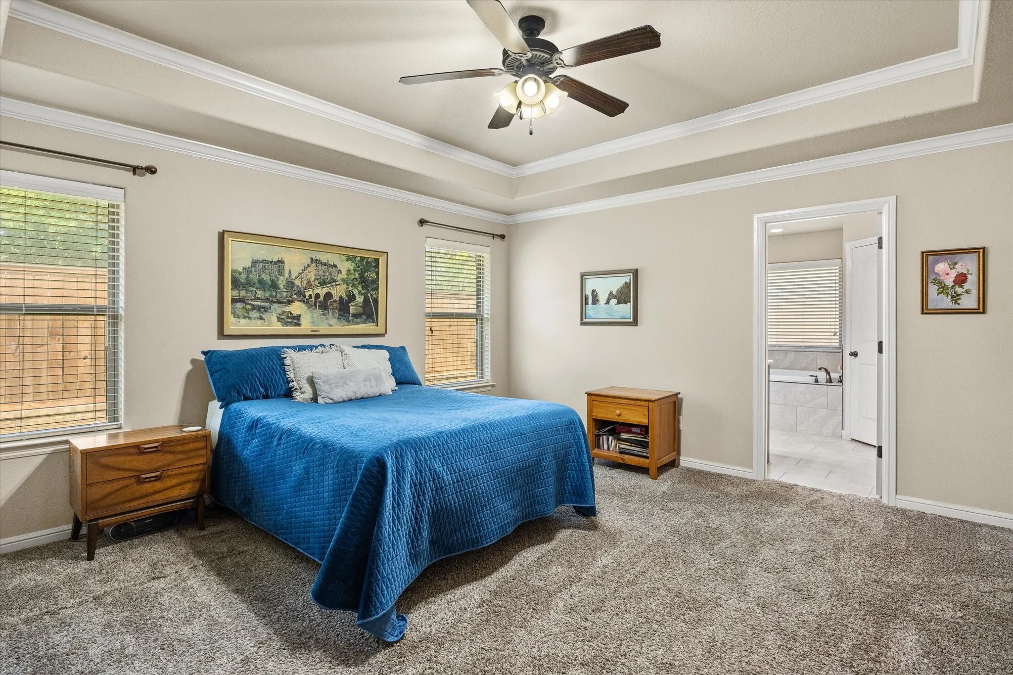 Tray ceiling with crown molding, ceiling fan, two windows with blinds, carpeted flooring, and ensuite bath