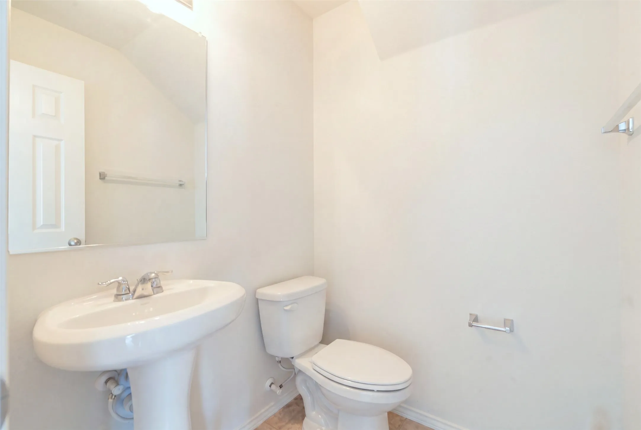 Clean, classic bathroom with a pedestal sink, chrome fixtures, wall-mounted mirror, white toilet, and tile flooring
