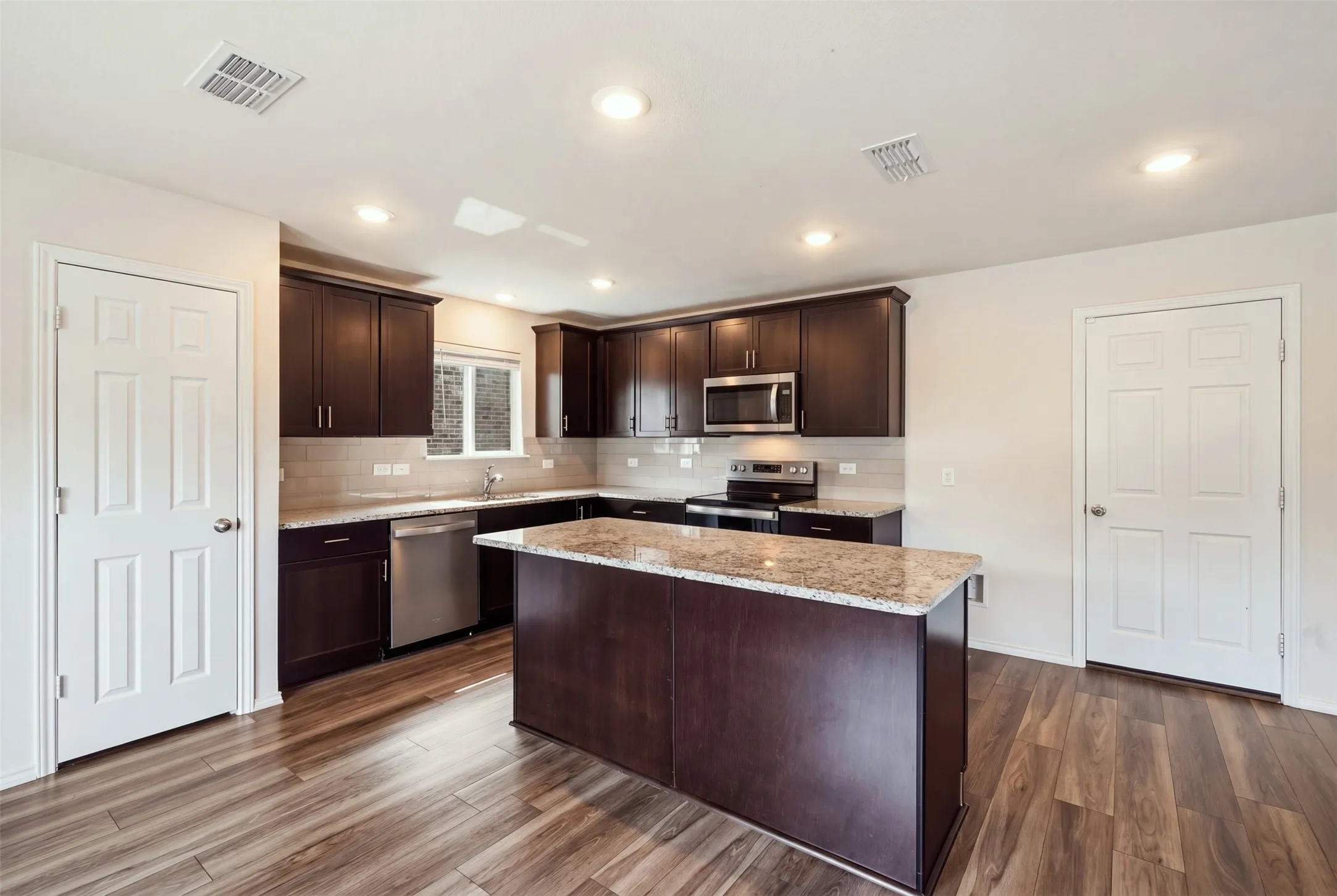 Stylish kitchen with warm wood-look floors, dark cabinetry, light countertops, stainless steel appliances, and a central island