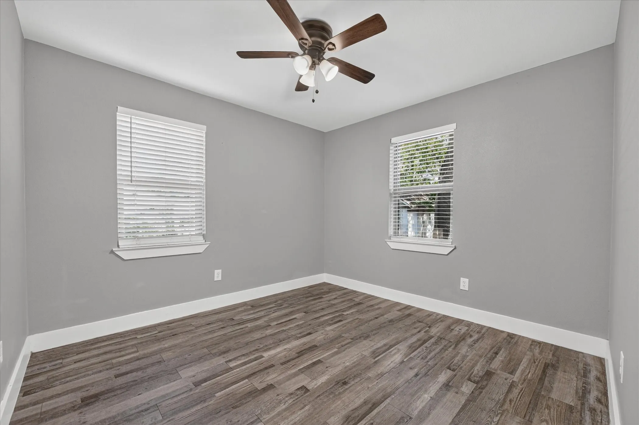 Light gray walls with white trim, wood-finish flooring, two windows with blinds, and a ceiling fan
