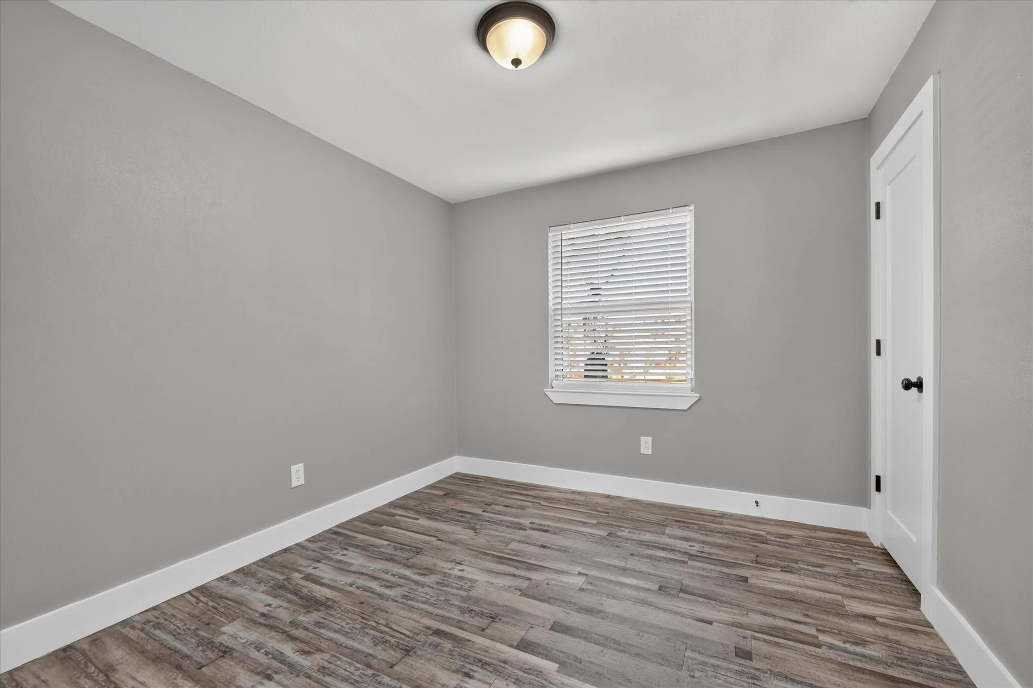 Neutral tone walls, wood-finish flooring, white trim, window with blinds, and a paneled door with dark hardware