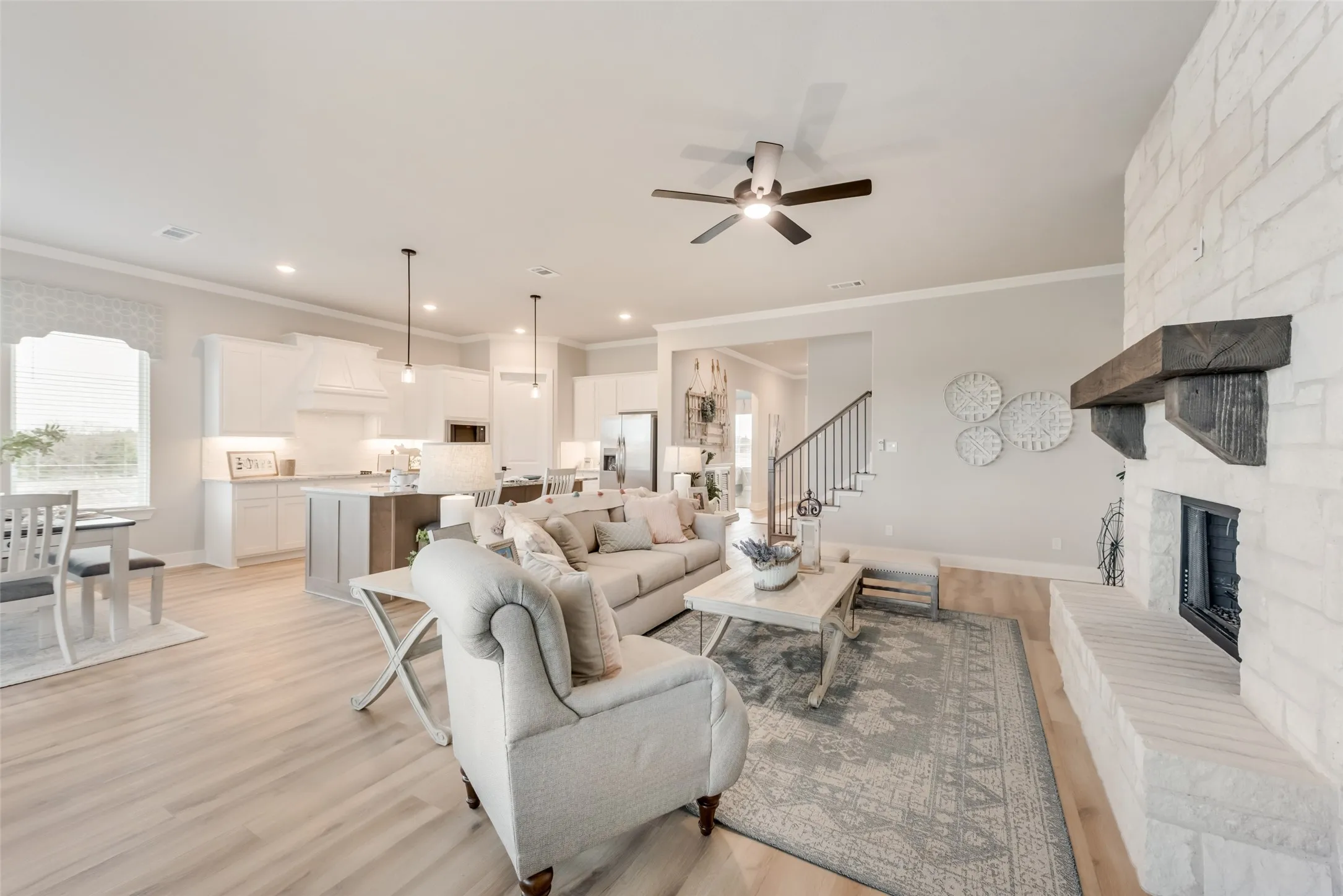 Spacious living area featuring wood-finish flooring, a white stone fireplace with a substantial wood mantel, and an open-concept kitchen with white cabinetry and pendant lighting