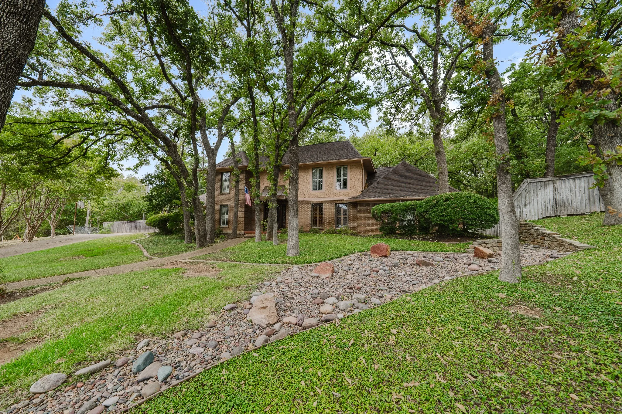 Brick exterior with multiple windows, mature trees, a manicured lawn, and a stone-lined dry creek bed