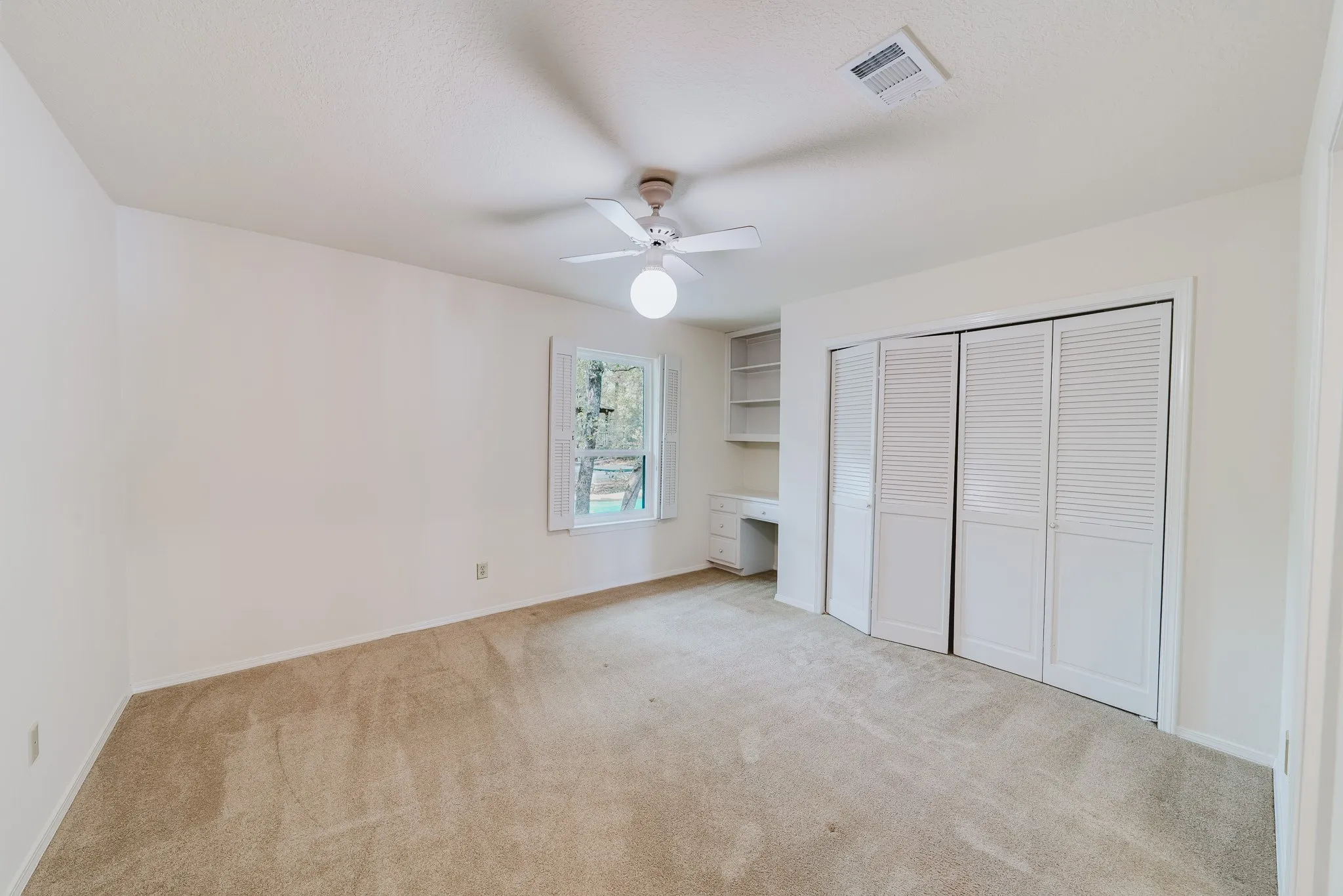Neutral tone carpeting, built-in desk with shelving, window with natural light, louvered bi-fold closet doors, and ceiling fan
