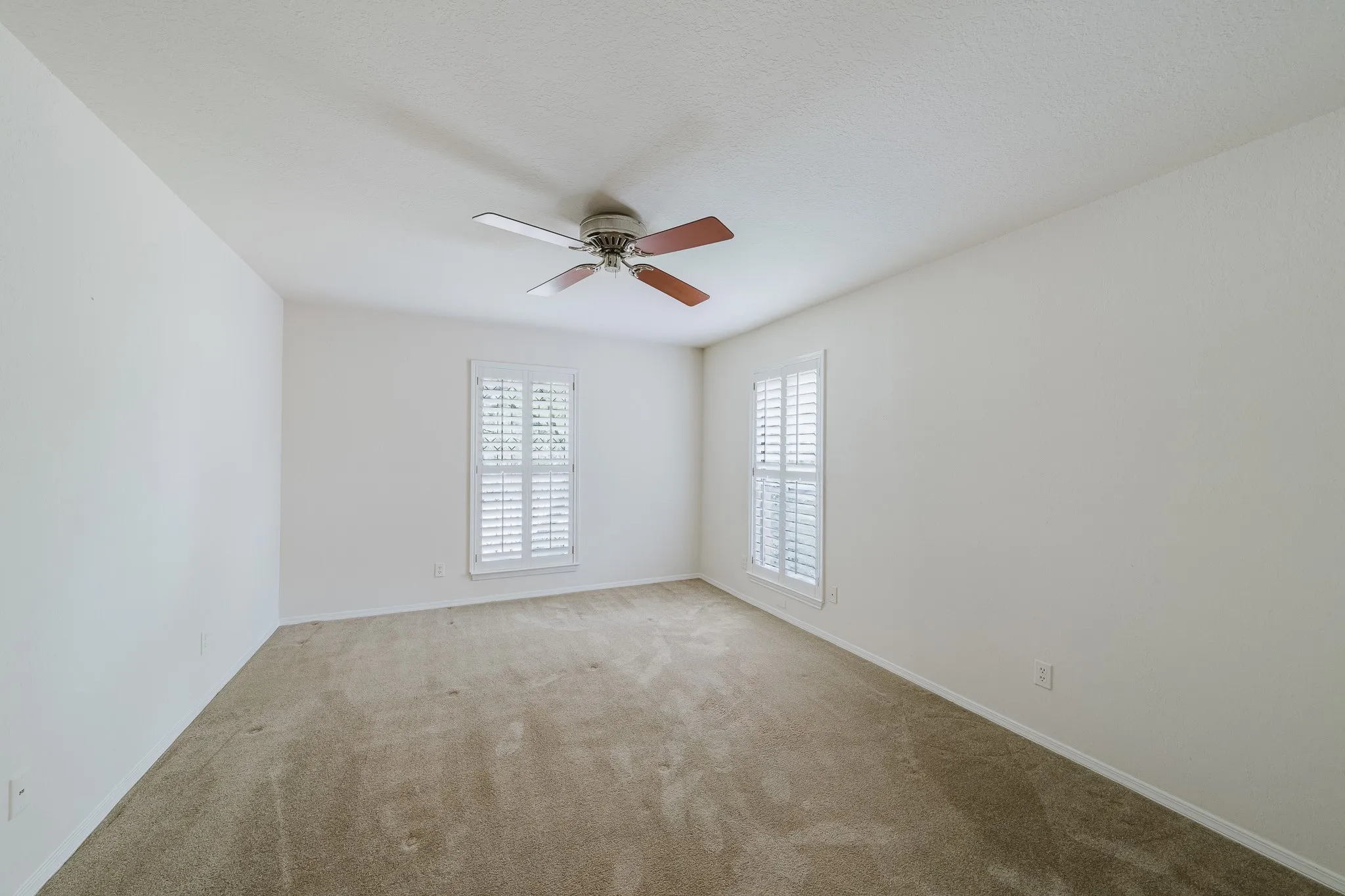 Neutral-toned wall-to-wall carpeting, white painted walls, two windows with white plantation shutters, and a ceiling fan with wood-finish blades