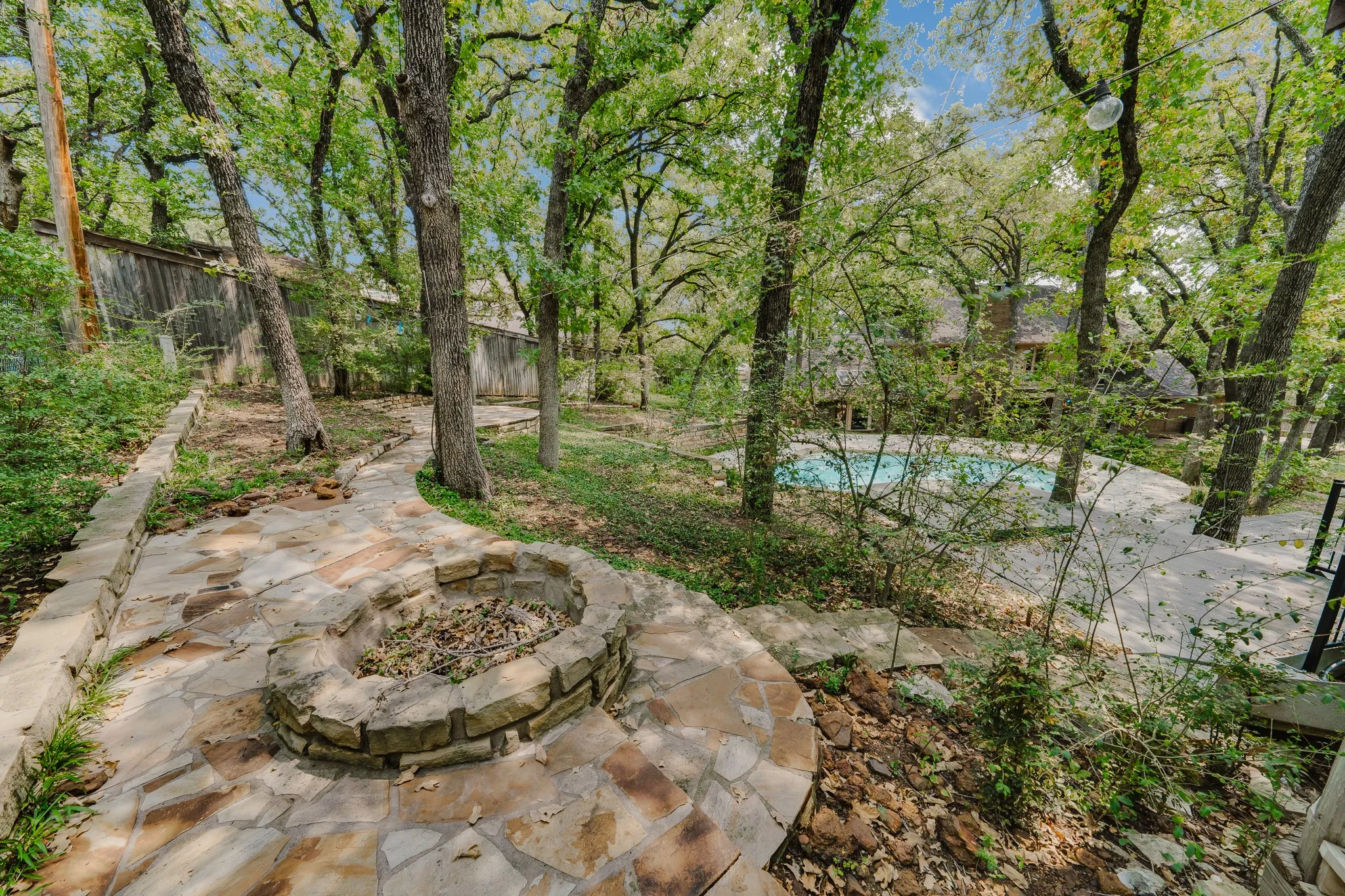 Stone paver pathway leading to a built-in stone fire pit, surrounded by mature trees, with a visible swimming pool in the background