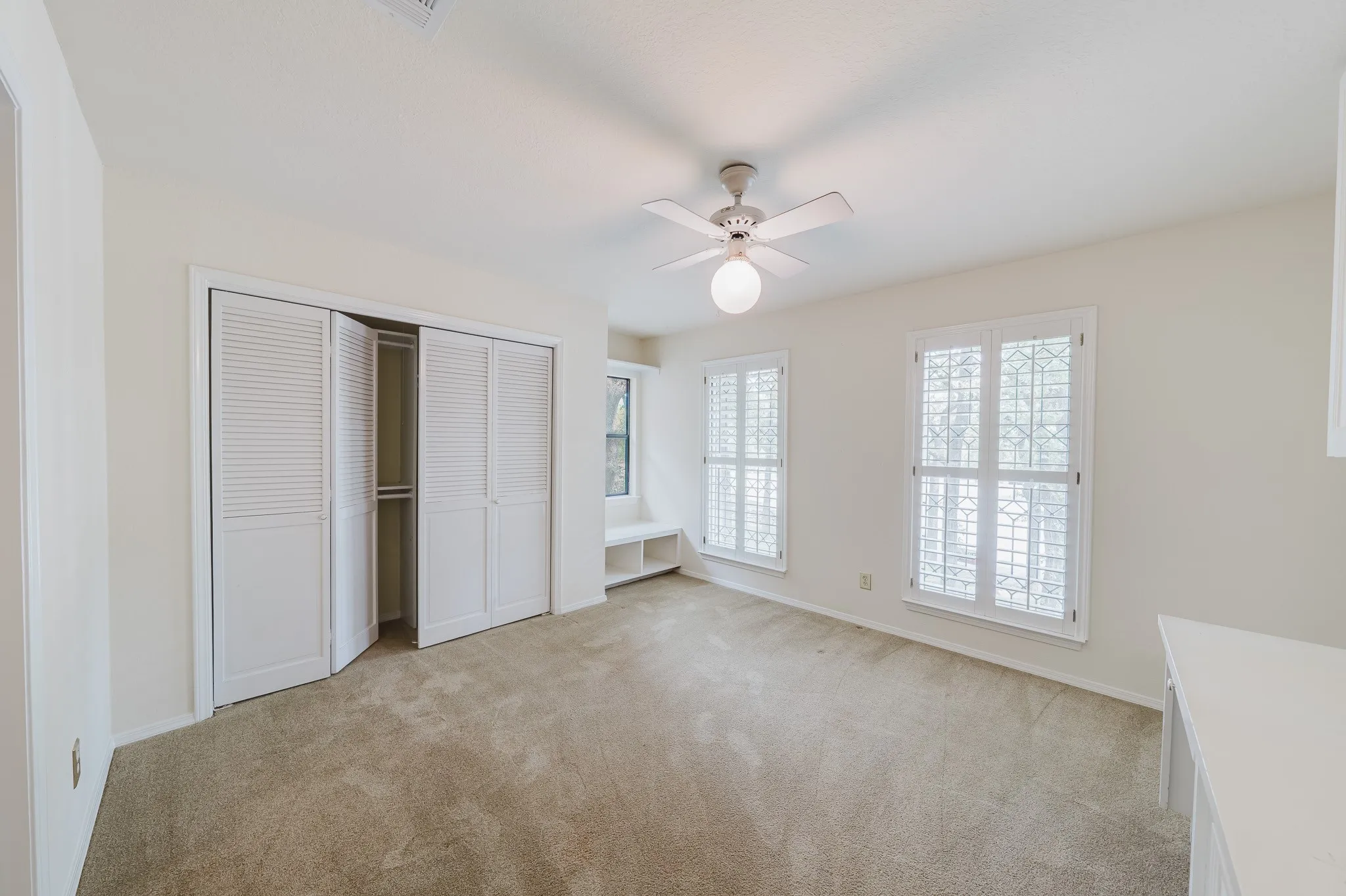 Plush carpet flooring, louvered bi-fold closet doors, ceiling fan with integrated lighting, built-in window seat, and white plantation shutters