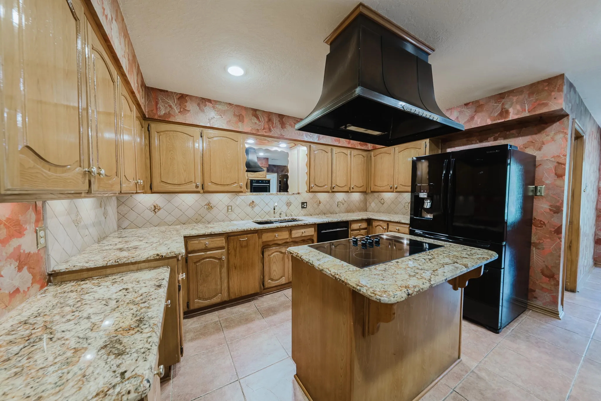 U-shaped kitchen layout featuring wood cabinetry, light granite countertops, a tiled backsplash, and a central island with an integrated cooktop