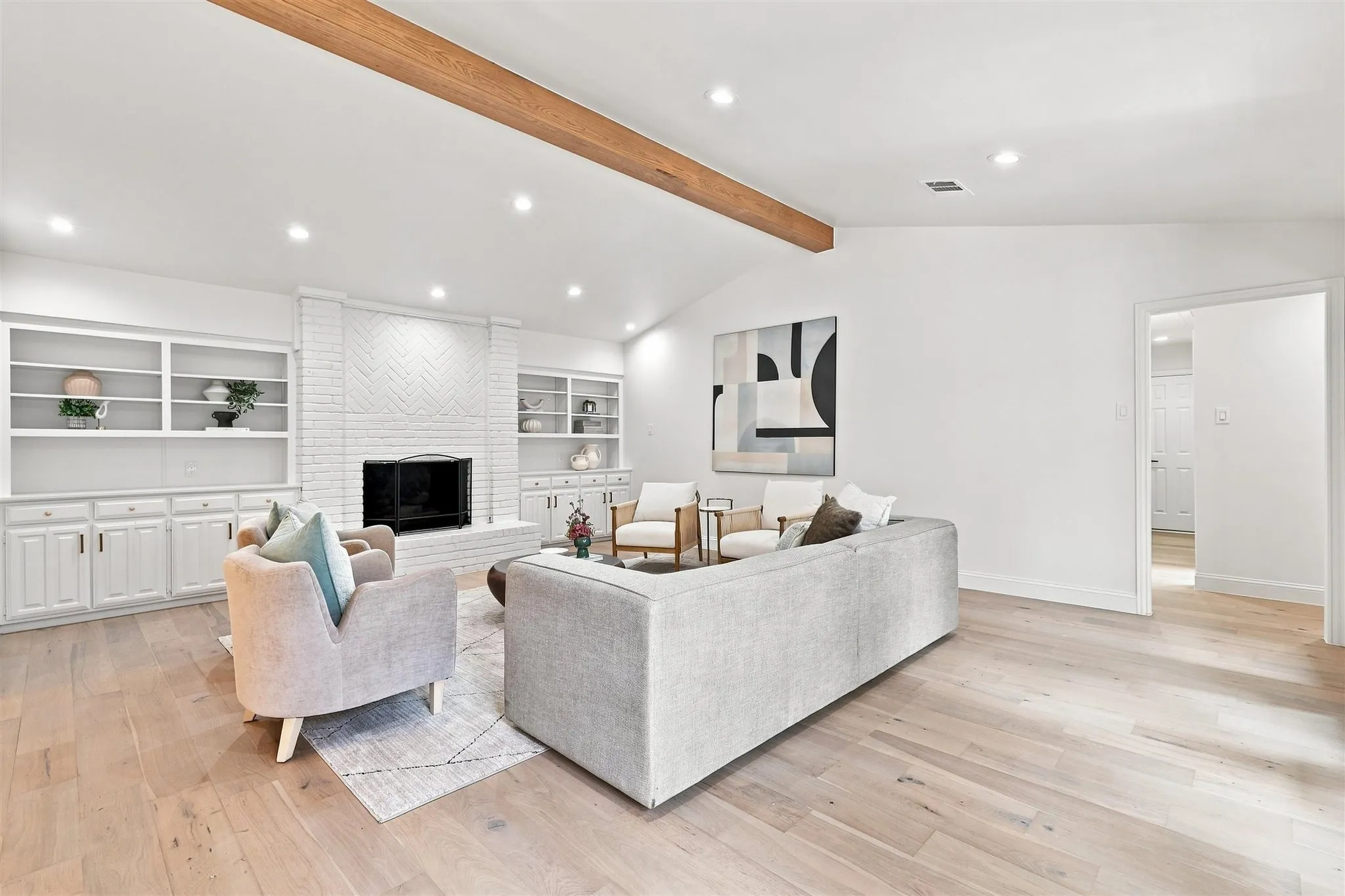 Vaulted ceiling with an exposed wood beam, recessed lighting, wood-finish flooring, white painted brick fireplace, and built-in cabinetry with open shelving
