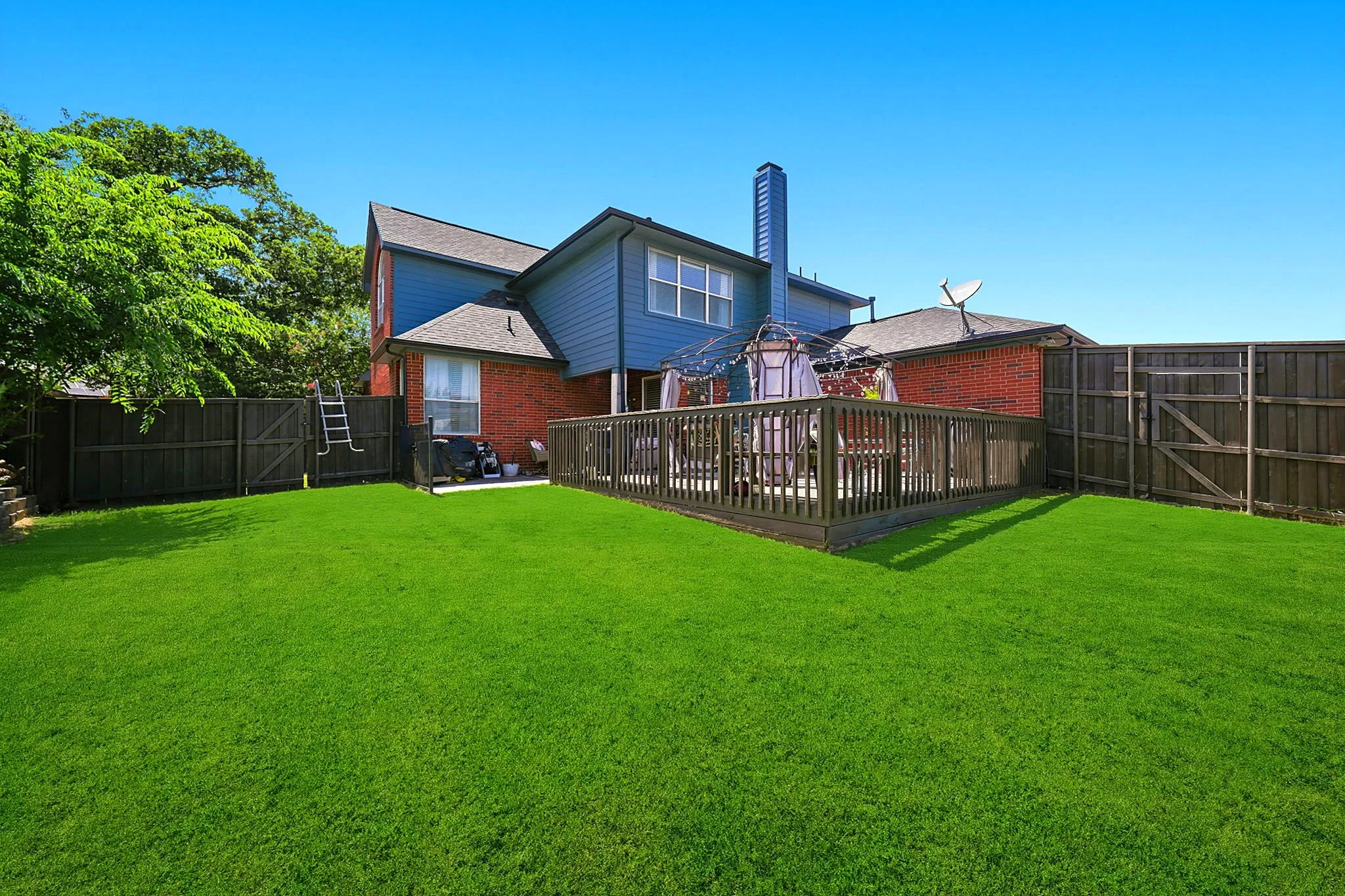 Expansive green lawn, wood fence, multi-level wood deck, brick and siding exterior, and mature trees