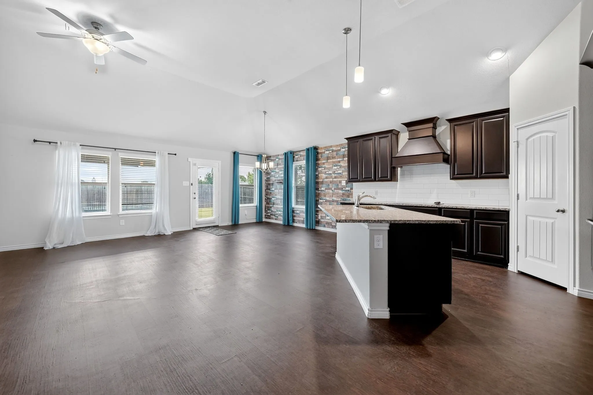 Vaulted ceilings, wood-finish flooring, kitchen island with sink, dark wood cabinetry, and a stacked stone accent wall