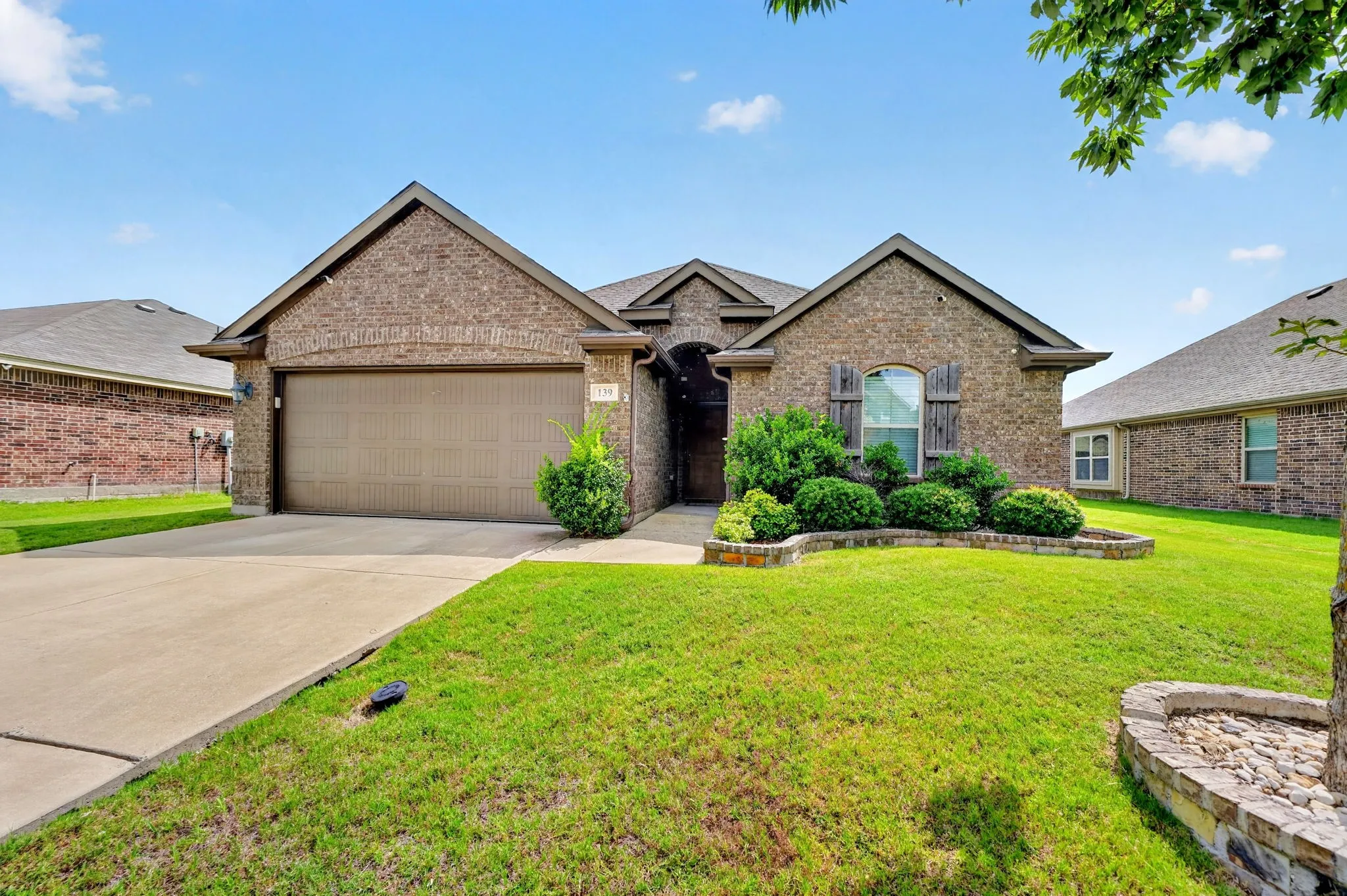 Brick exterior with gabled roof lines, attached garage, concrete driveway, and landscaped front yard with stone-edged planters