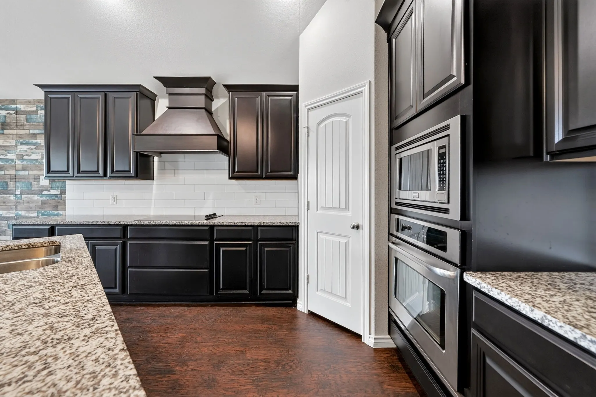 Dark-stained cabinetry, stainless steel built-in appliances, light granite countertops, white subway tile backsplash, and wood-finish flooring