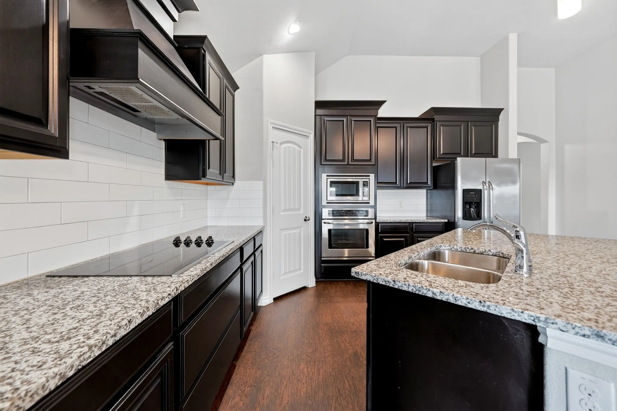 Kitchen featuring dark wood-finish cabinetry, light speckled countertops, a built-in cooktop, integrated stainless steel appliances, and a double basin sink