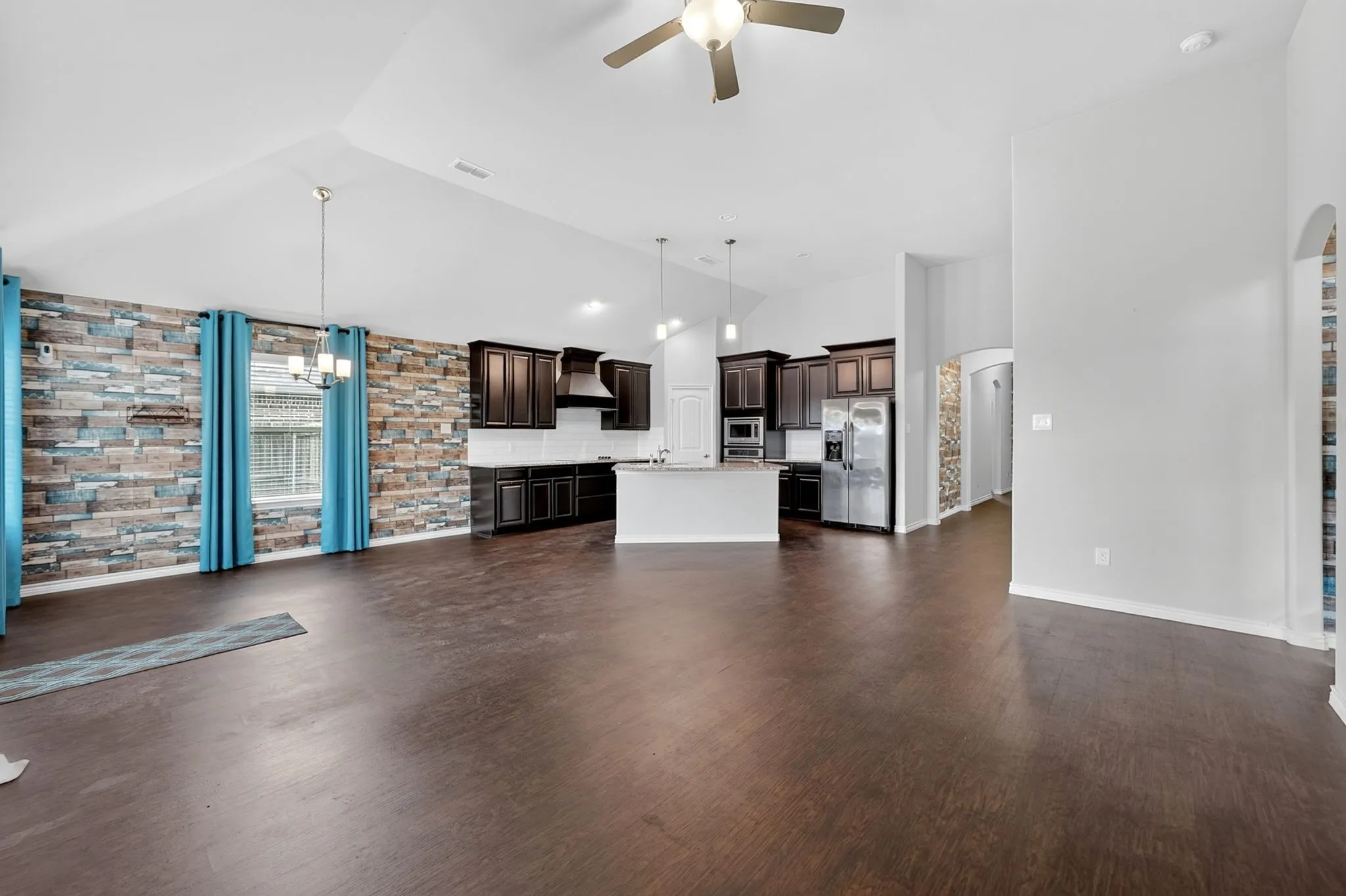 Open-concept living space featuring soaring vaulted ceilings with ceiling fan, wood-finish flooring throughout, and abundant natural light from large windows dressed in turquoise drapery