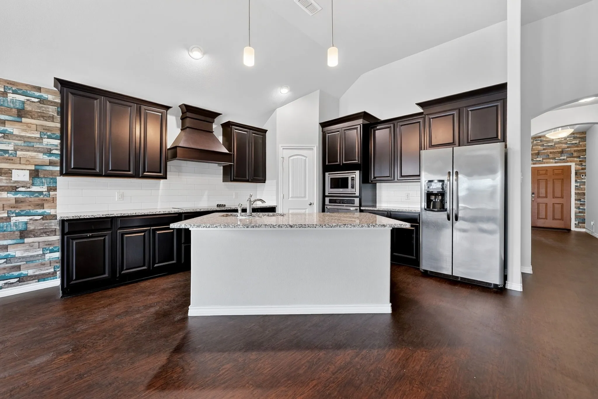 Kitchen featuring dark wood-finish cabinetry, built-in stainless steel appliances, a center island with a sink, white subway tile backsplash, and dark wood-finish flooring
