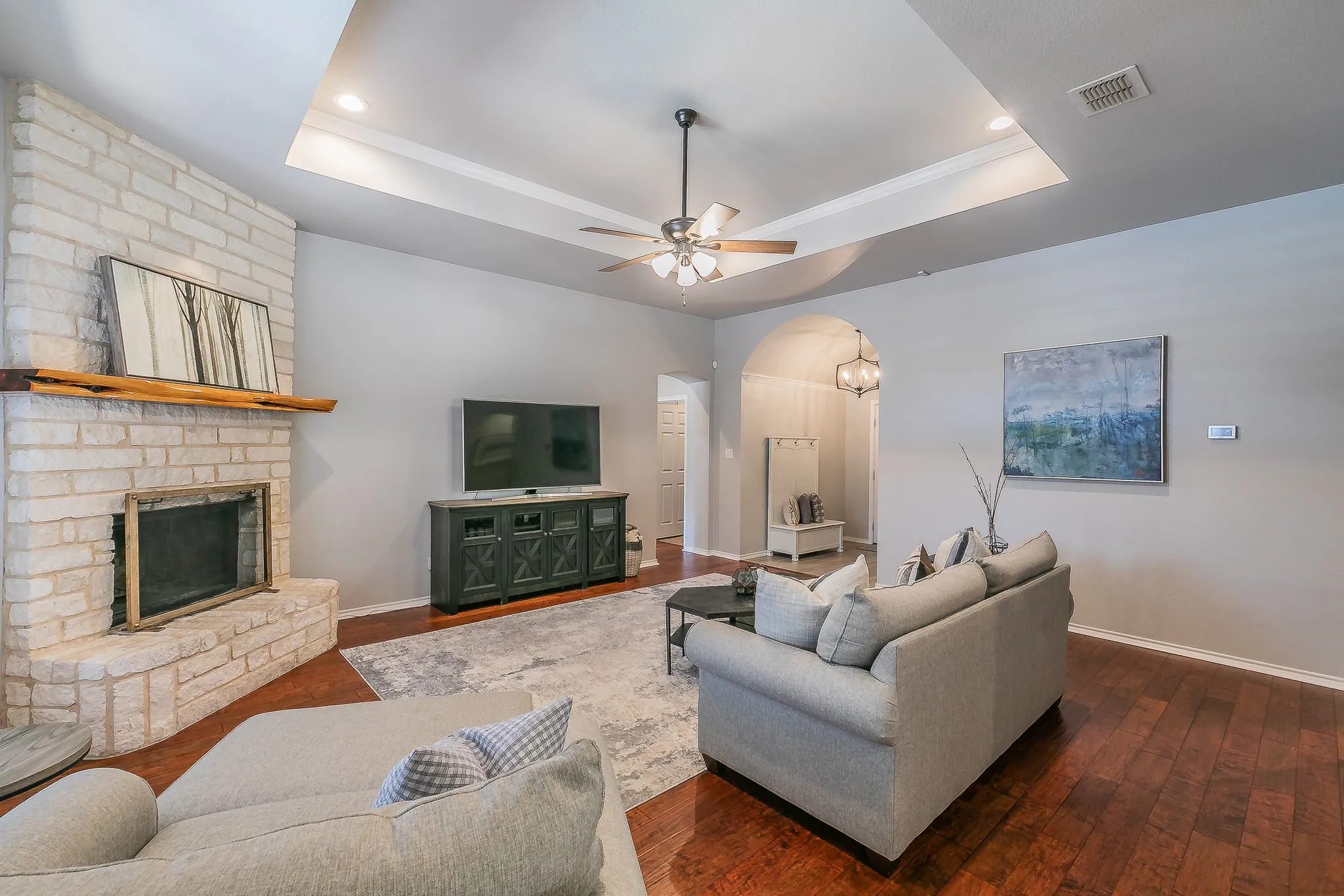 Tray ceiling with recessed lighting, wood-finish flooring, brick fireplace with wood mantel, ceiling fan, and an arched entry