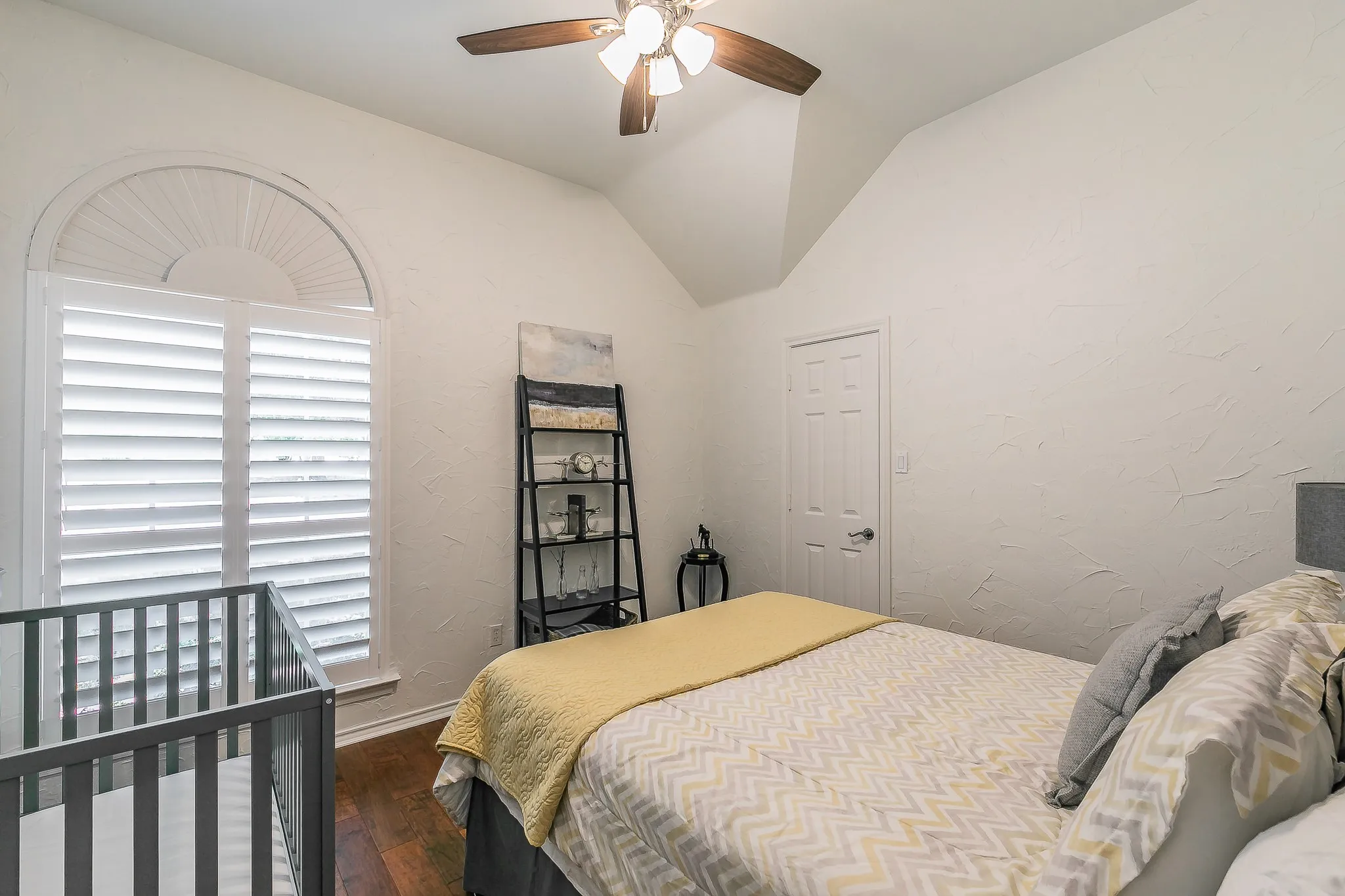 Arched window with plantation shutters, vaulted ceiling, ceiling fan with light fixture, wood-finish flooring, and white paneled door