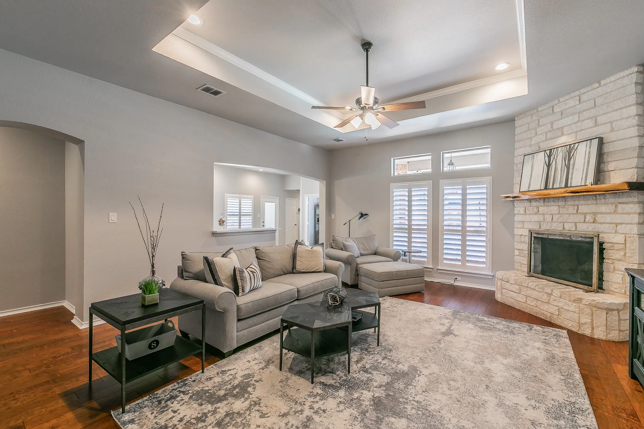Tray ceiling with recessed lighting and a ceiling fan, stone-surround fireplace with a wood mantel, wood-finish flooring, and white plantation shutters
