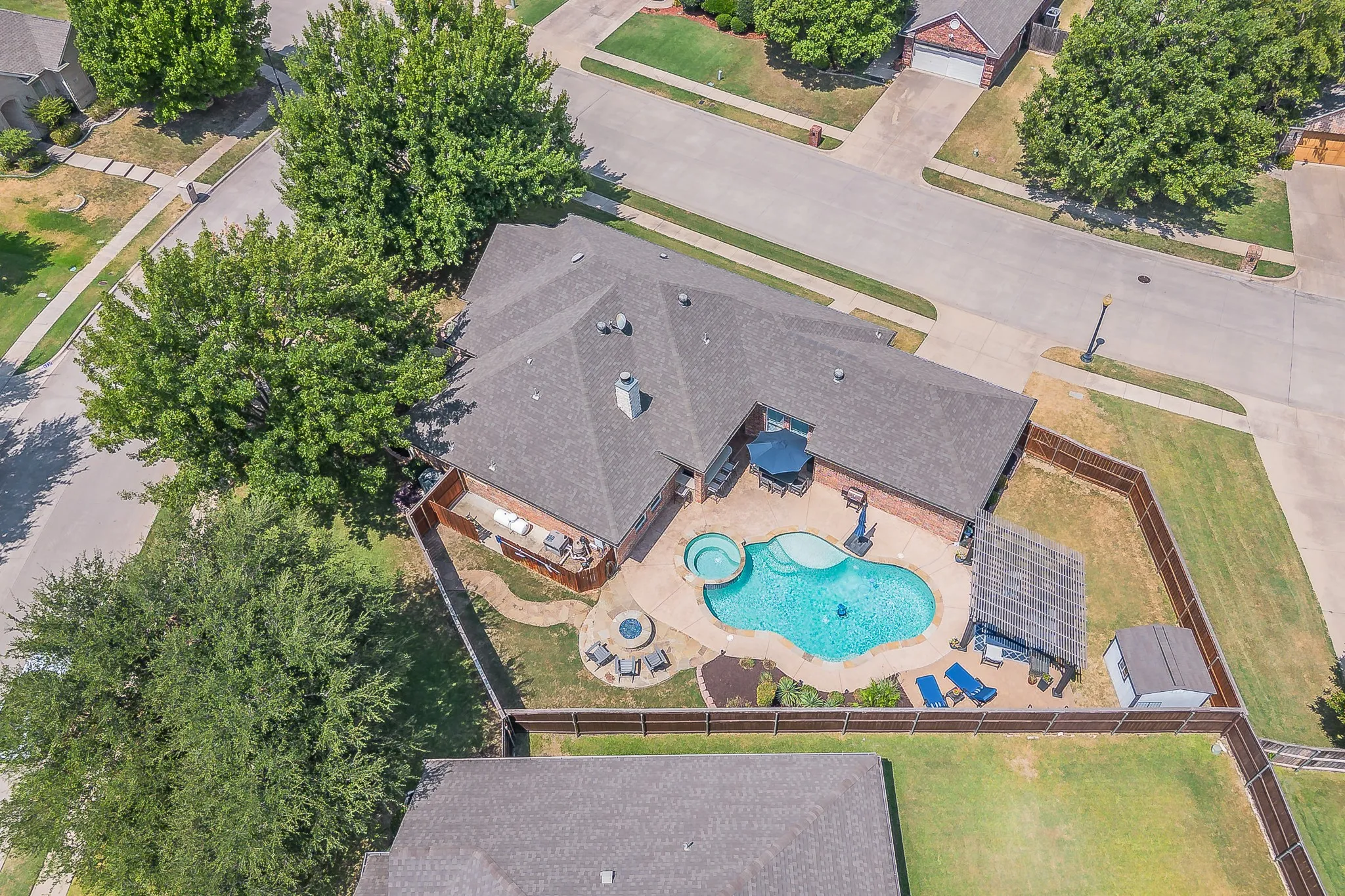 Shingled roofline over a brick exterior, featuring a freeform swimming pool with integrated spa, adjacent hardscaping, and a backyard with fencing