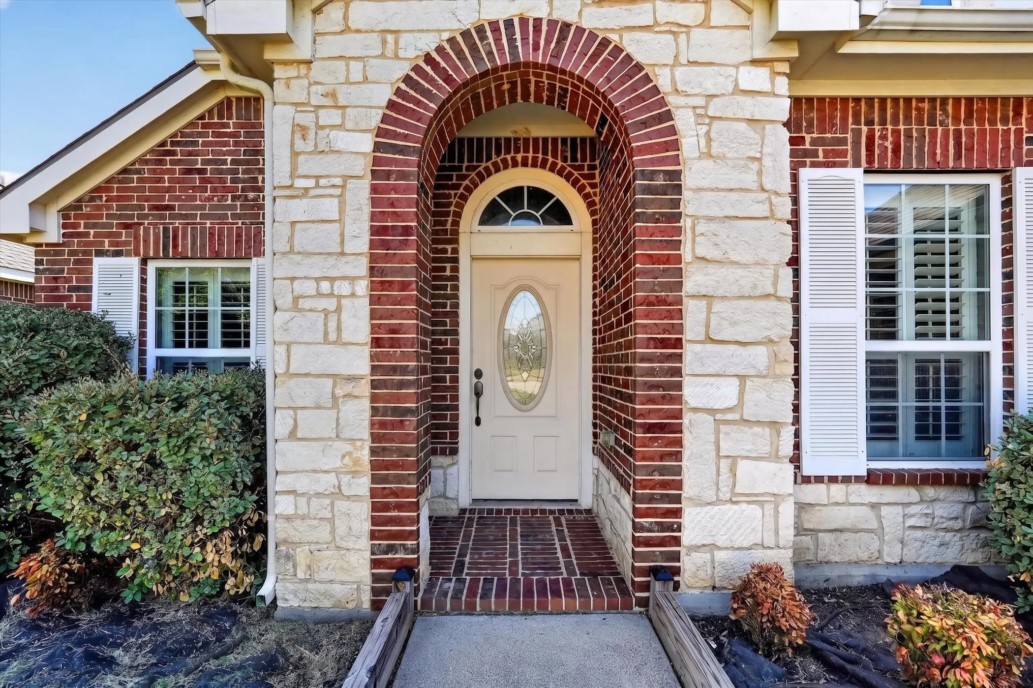 Brick and stone exterior with an arched entryway, decorative front door, and white louvered shutters