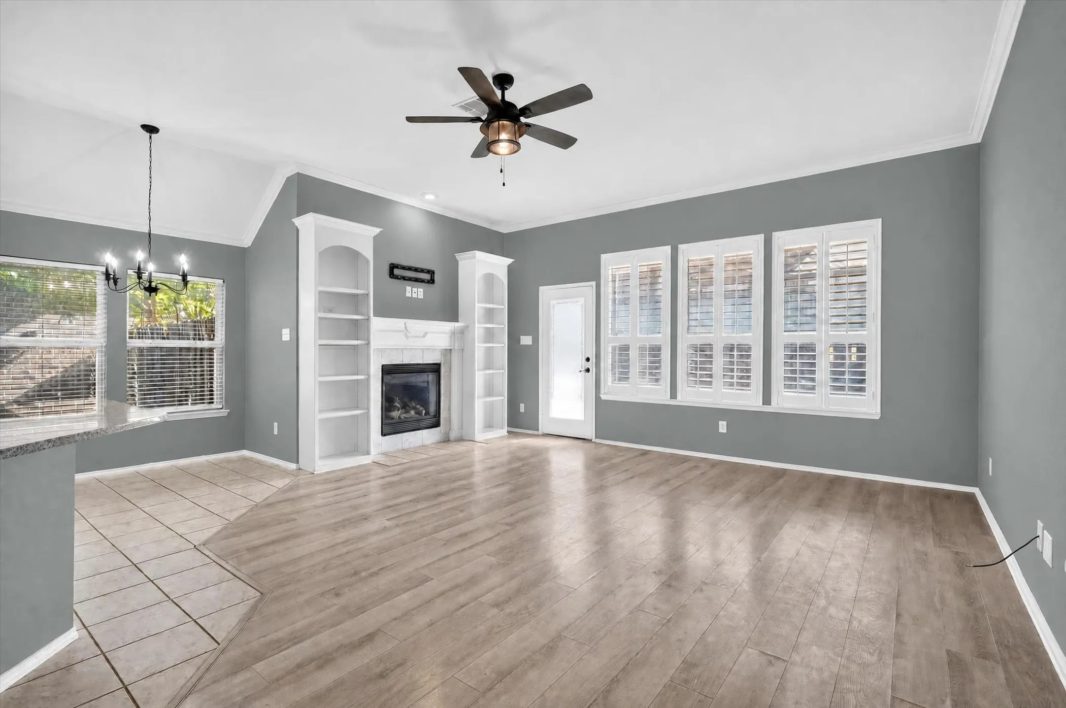 Spacious living area featuring a fireplace with built-in shelving, wood-finish flooring, and multiple windows with plantation shutters