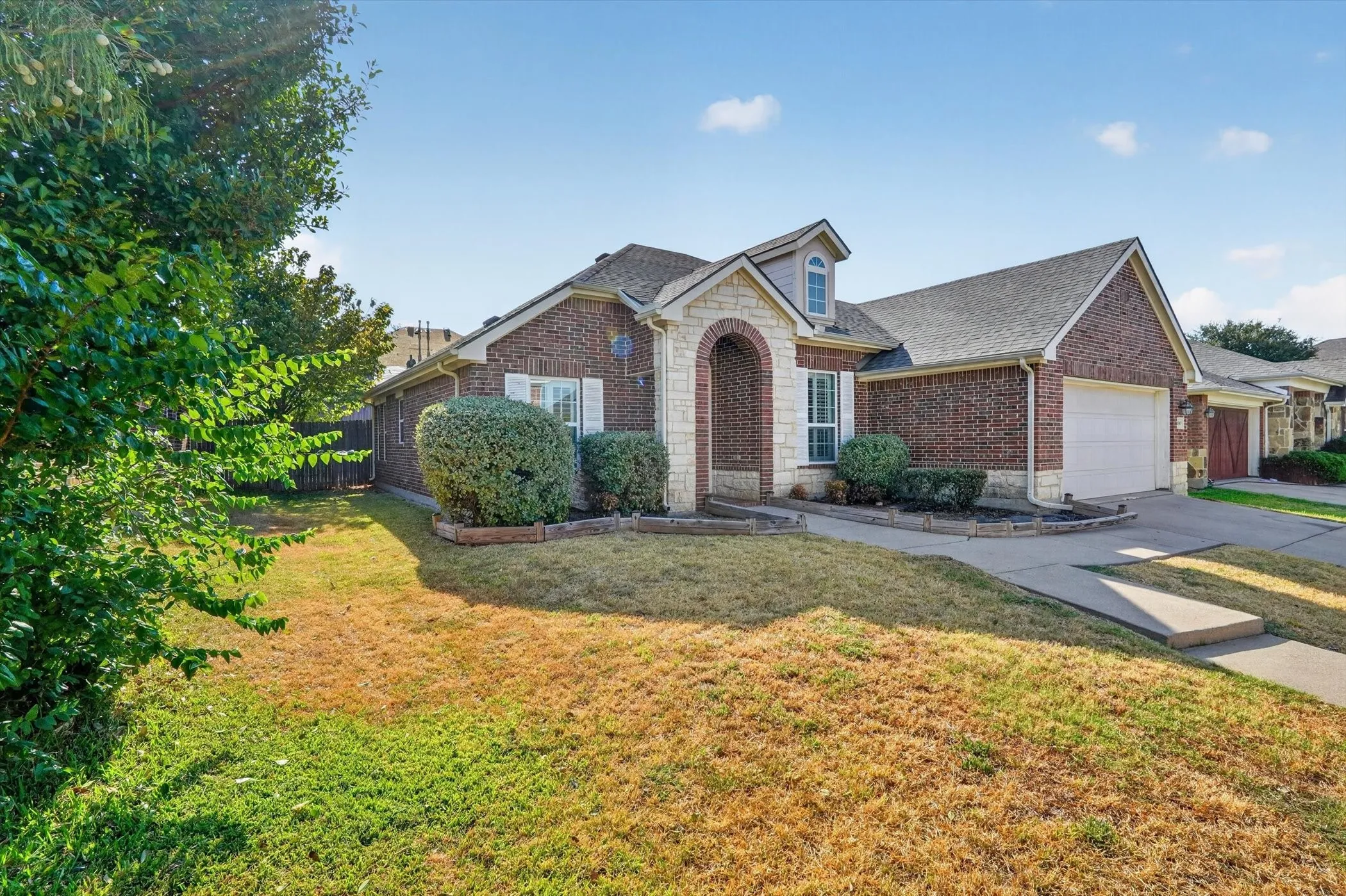 Brick exterior with stone accents, arched entry, dormer window, attached two-car garage, and established landscaping