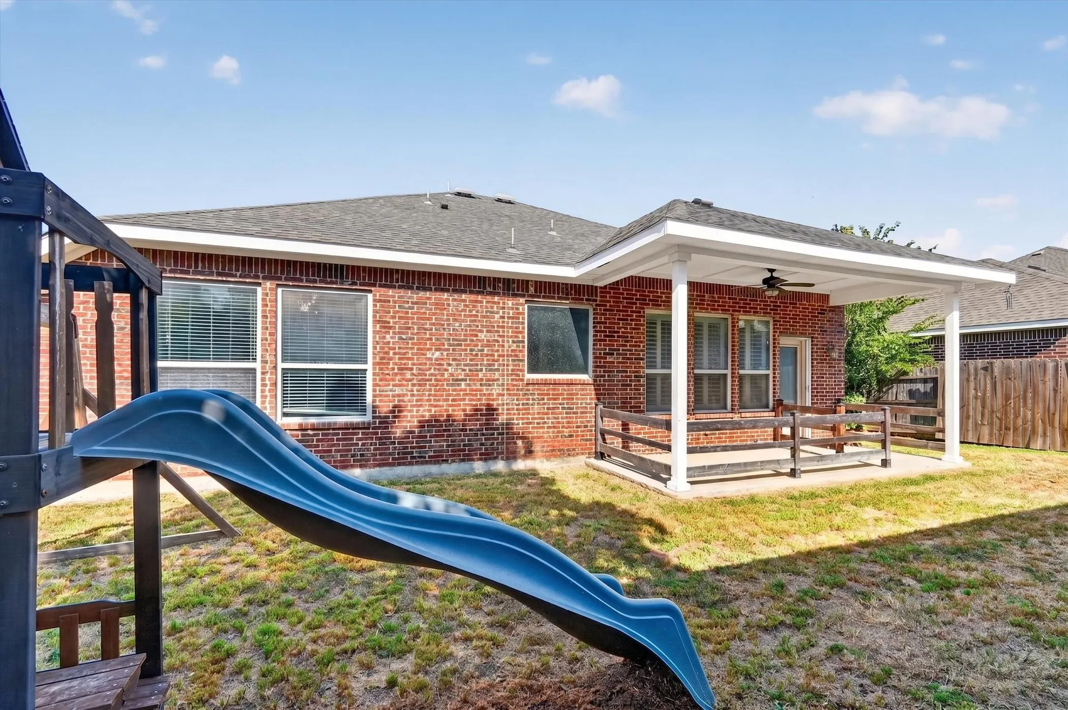 Brick exterior featuring multiple windows with blinds, a covered patio with a ceiling fan, and a wood-slat fence