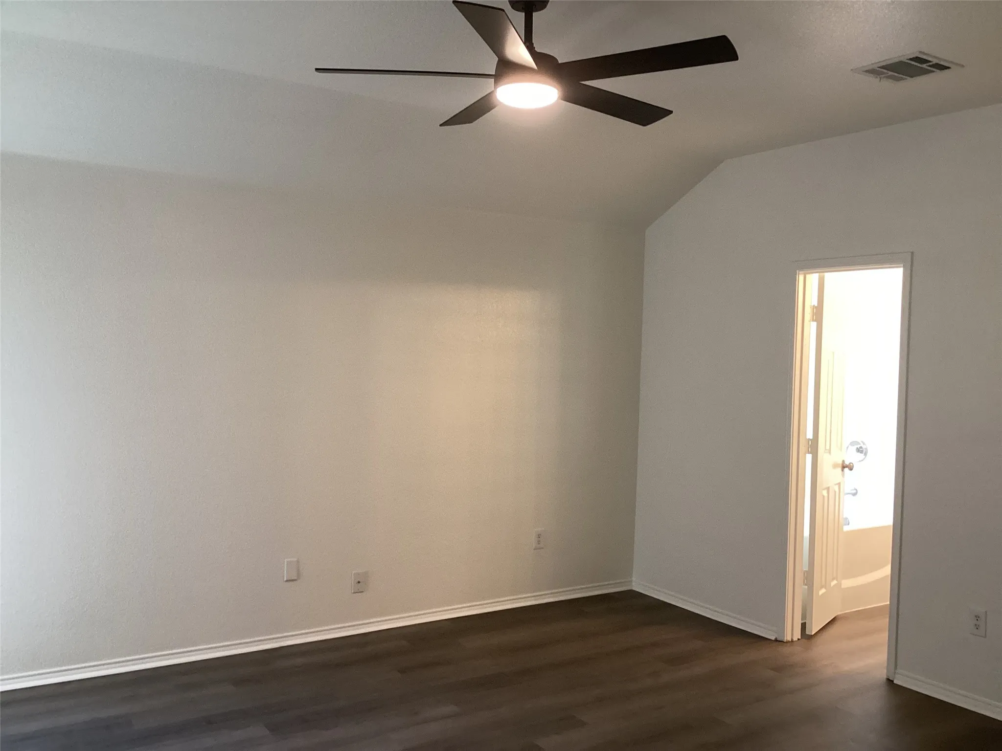 Vaulted ceiling, wood-finish flooring, ceiling fan with integrated lighting, crisp white walls, and a convenient en-suite bathroom entrance