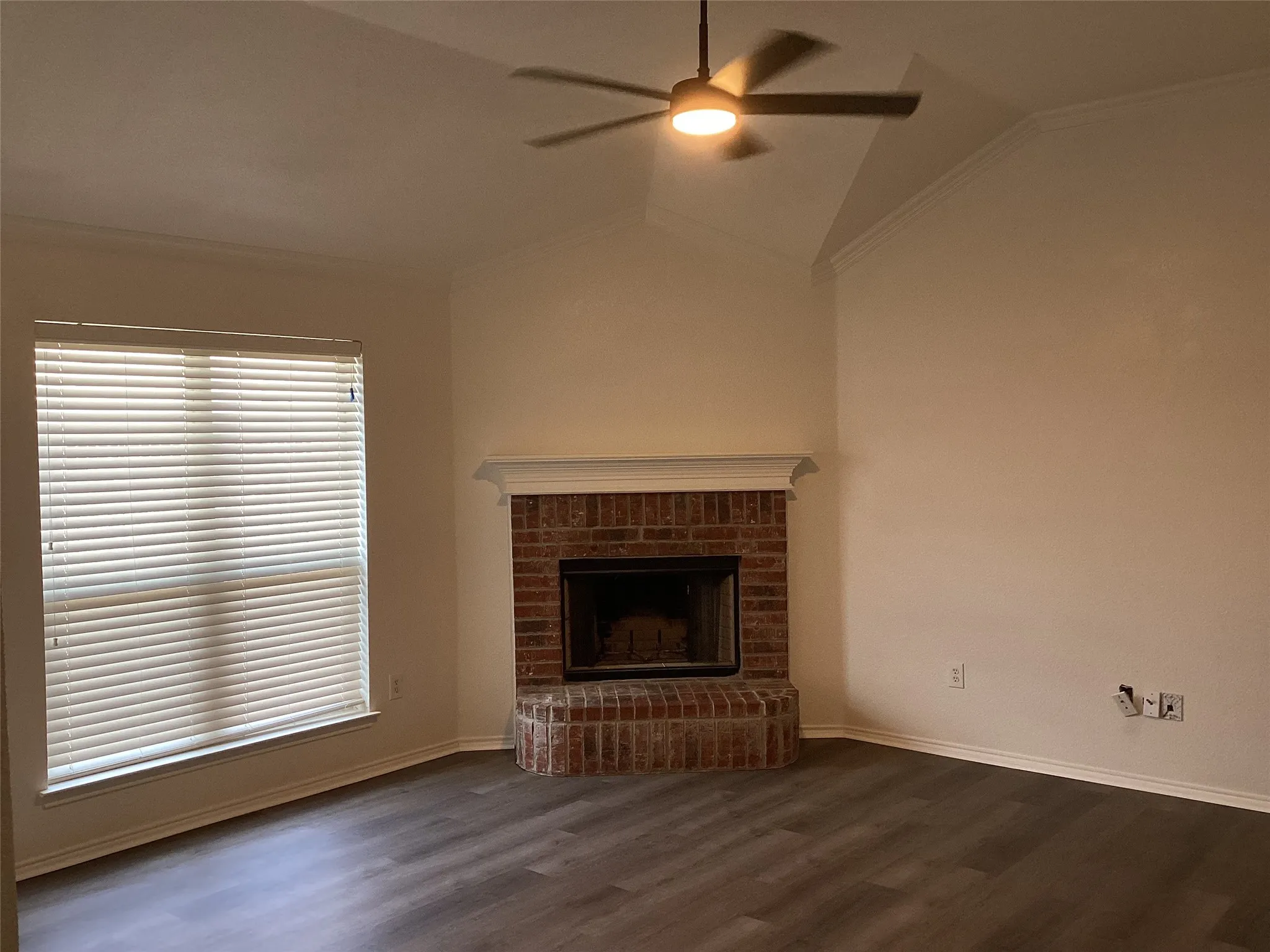 Vaulted ceiling with crown molding, brick fireplace with mantel, wood-finish flooring, and a ceiling fan with integrated lighting