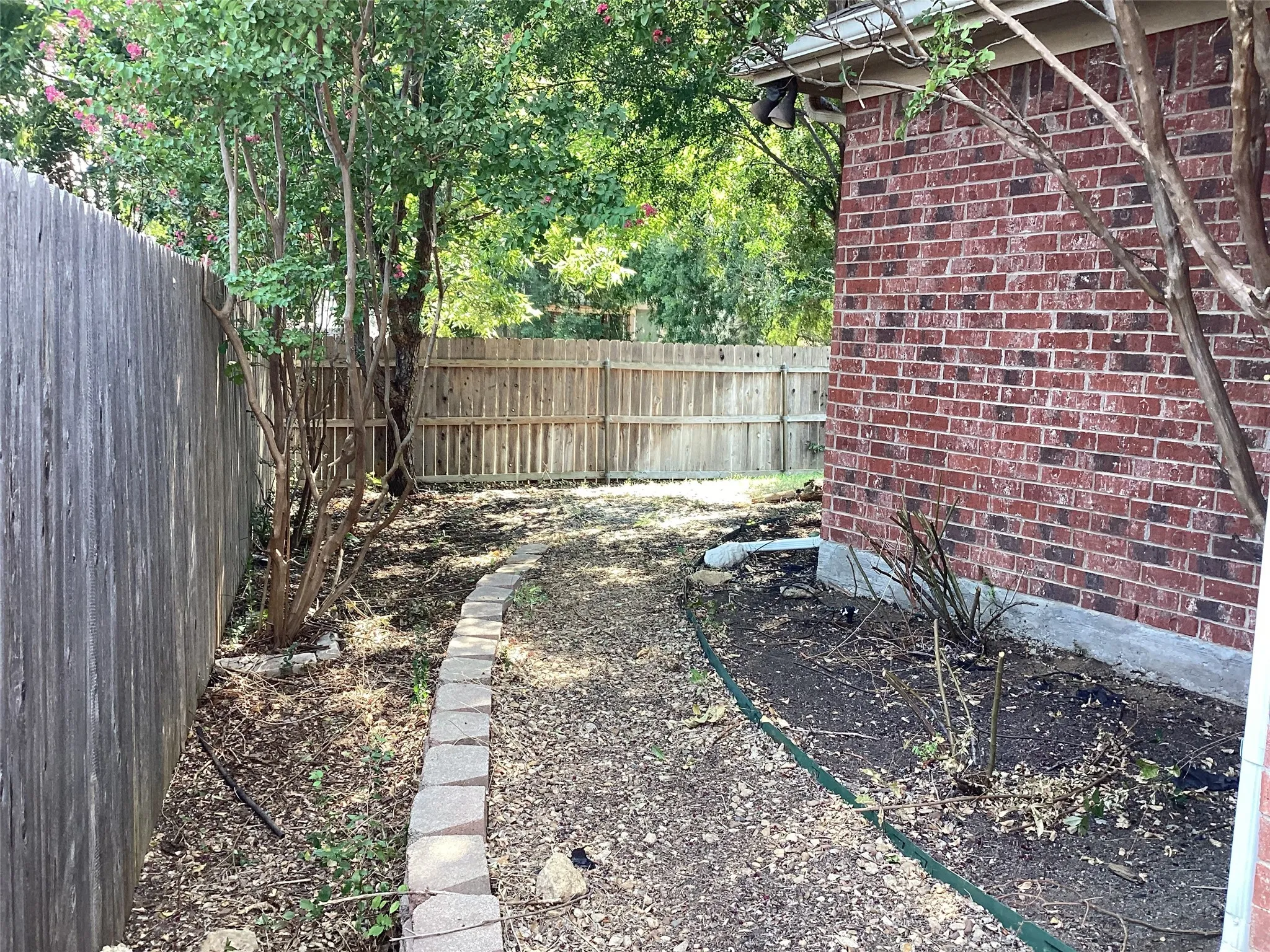 Red brick exterior, wood privacy fencing, stone-edged pathway, established trees, and planting beds