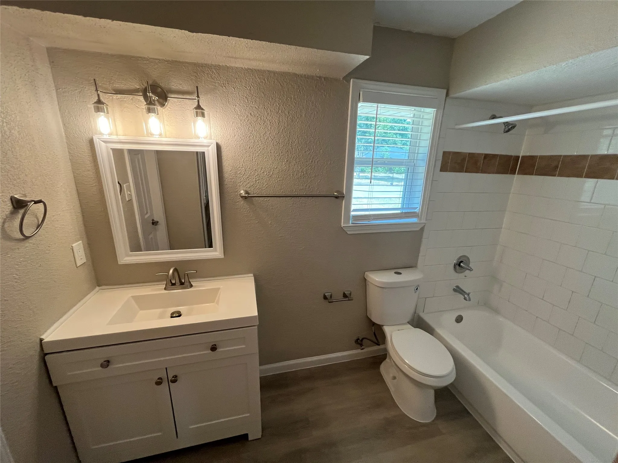 White vanity with integrated sink, brushed nickel fixtures, subway tile bath surround with accent band, and wood-finish flooring