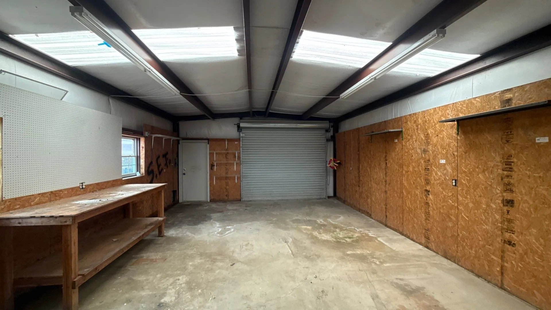 Spacious interior with exposed steel beams, multiple skylights, fluorescent lighting, a roll-up garage door, and a built-in workbench