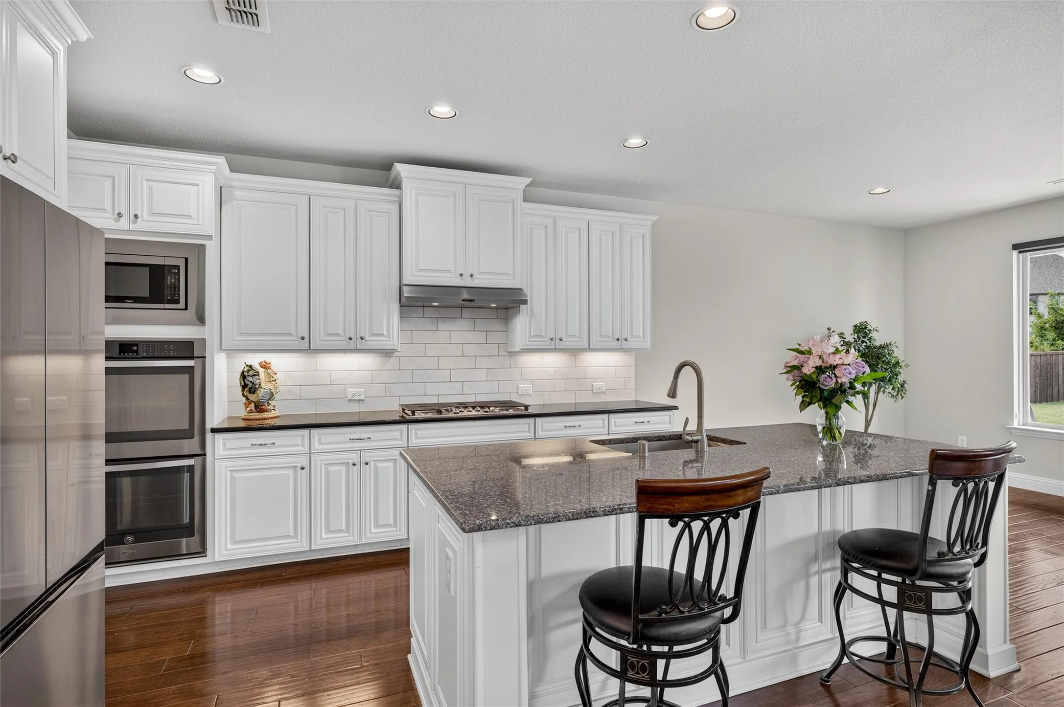 White shaker-style cabinetry, stainless steel appliances, dark finish countertops, wood-finish flooring, and recessed lighting