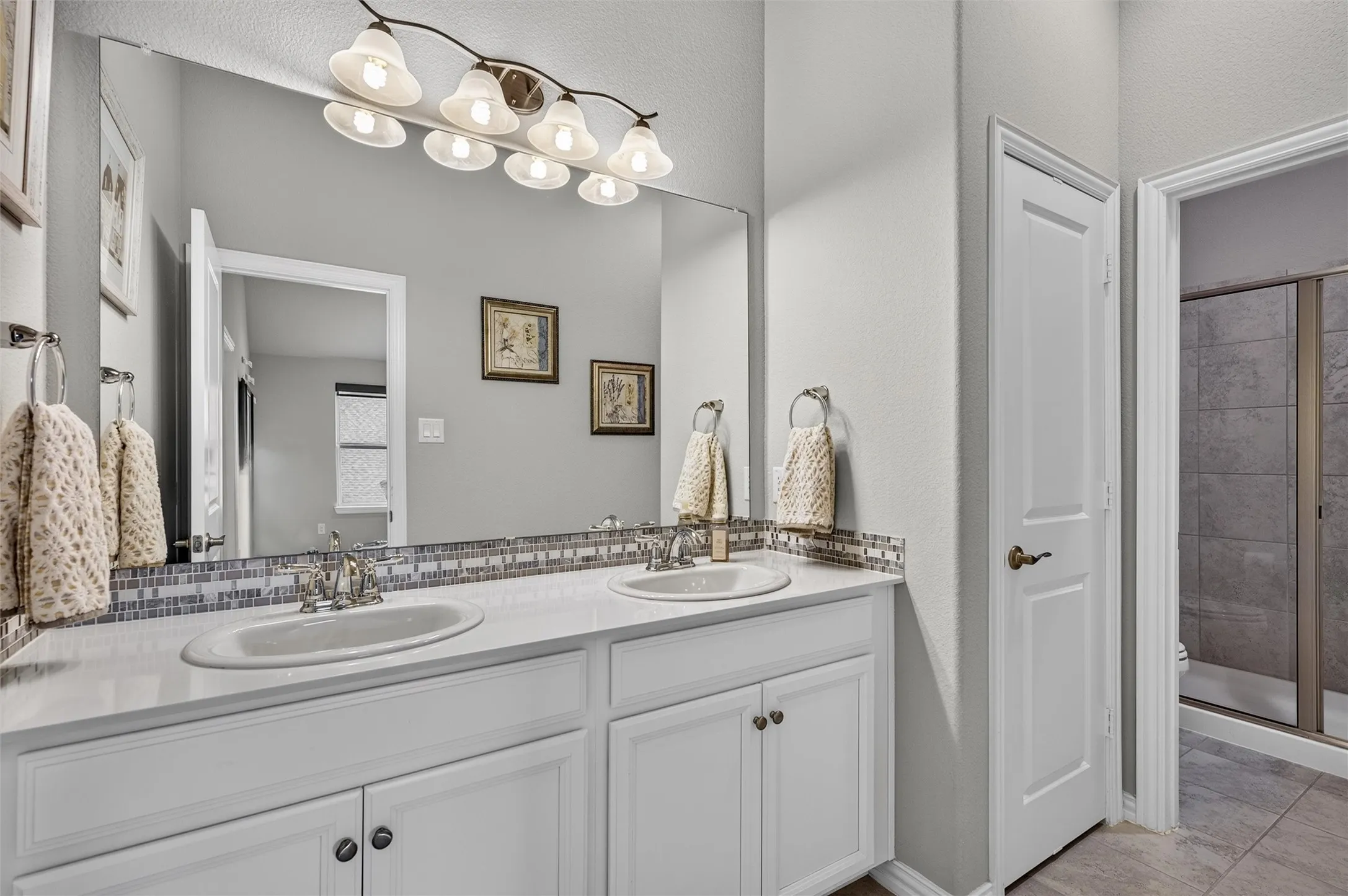 Dual undermount sinks, white cabinetry with dark hardware, full-width mirror, mosaic tile backsplash, and a separate walk-in shower with glass enclosure