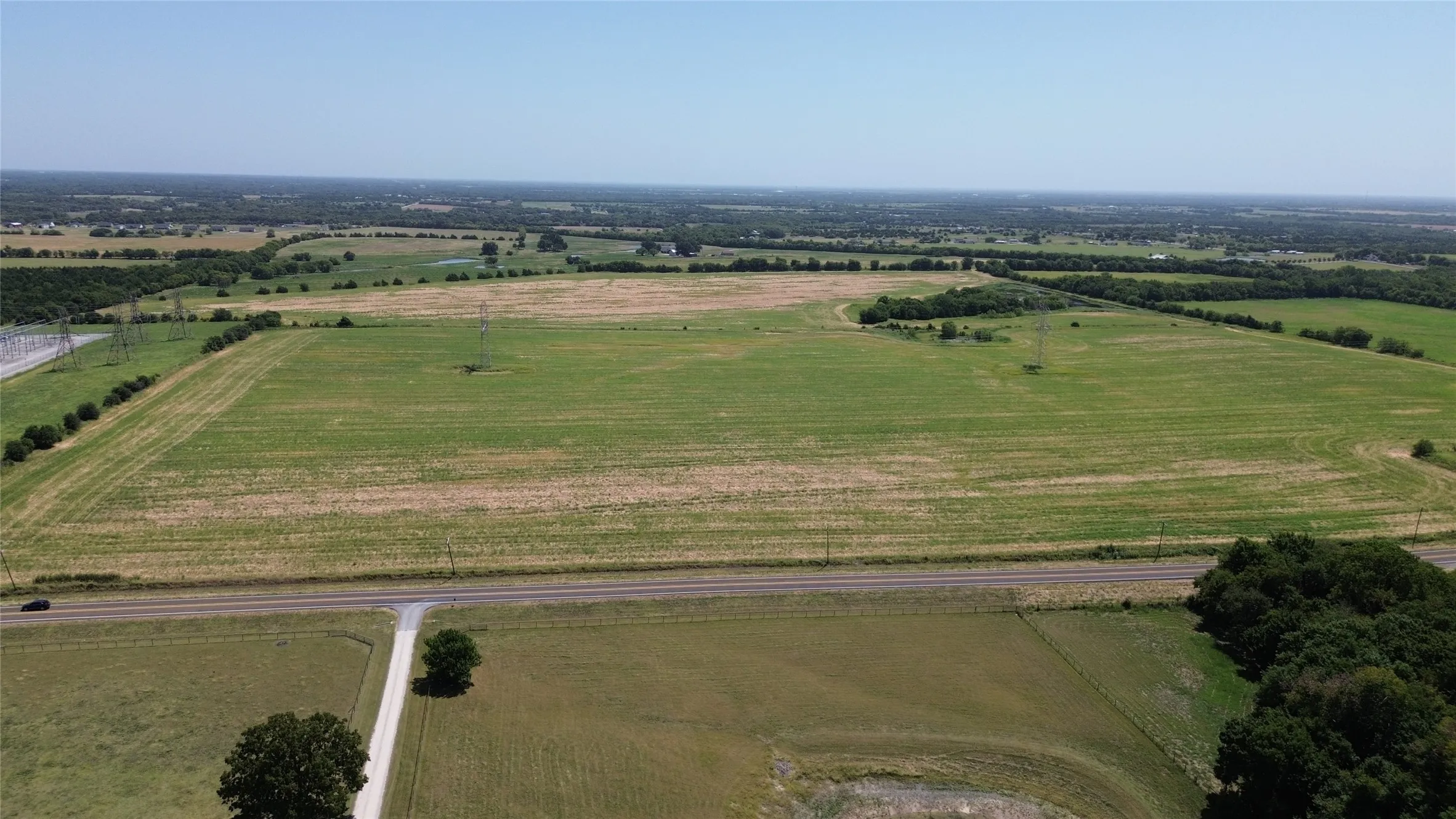 Expansive land parcel with clear skies, featuring a paved road, scattered trees, and visible power lines
