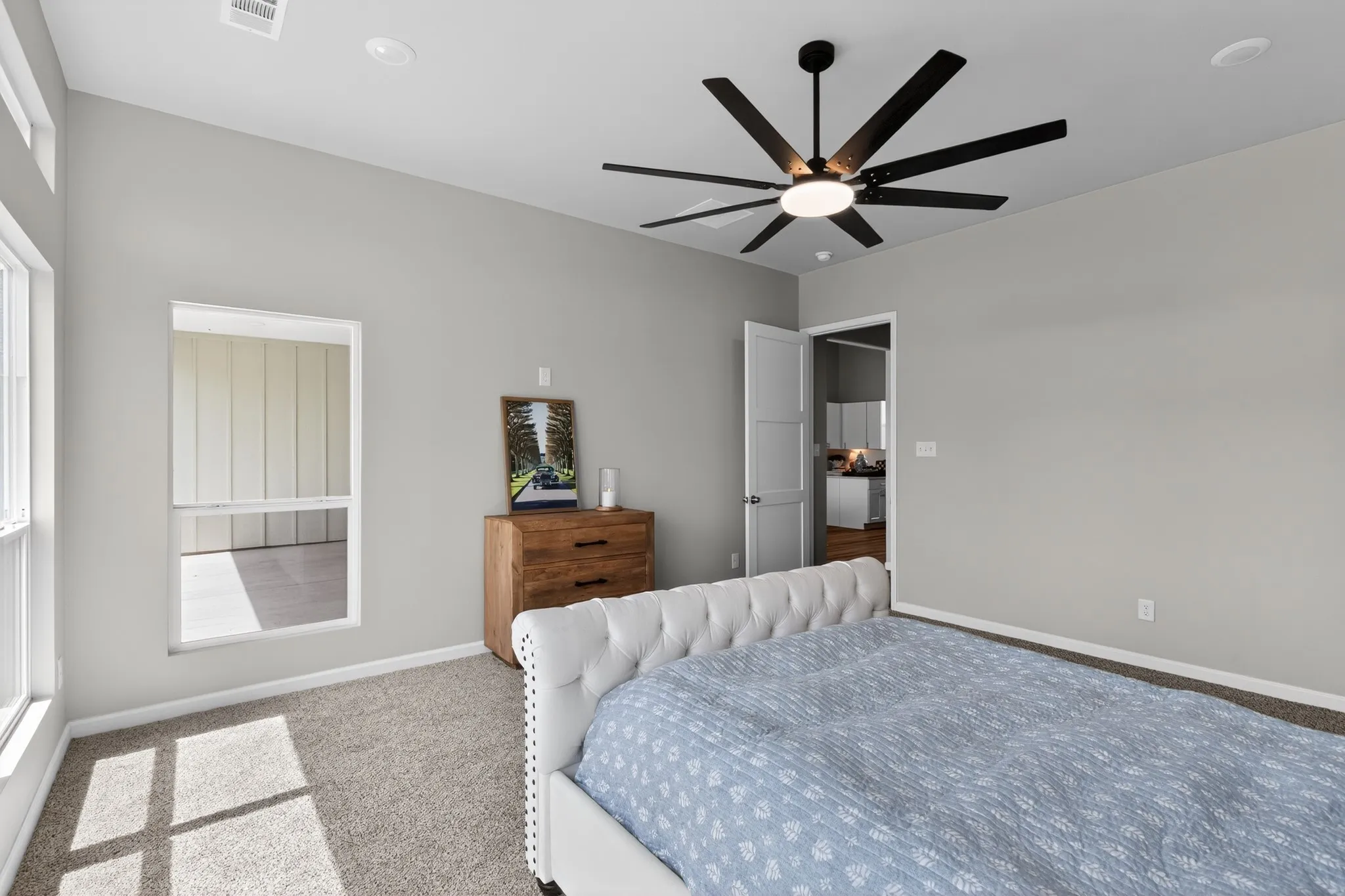 Spacious room featuring neutral carpeting, light gray walls, a modern black ceiling fan, and multiple windows providing natural light