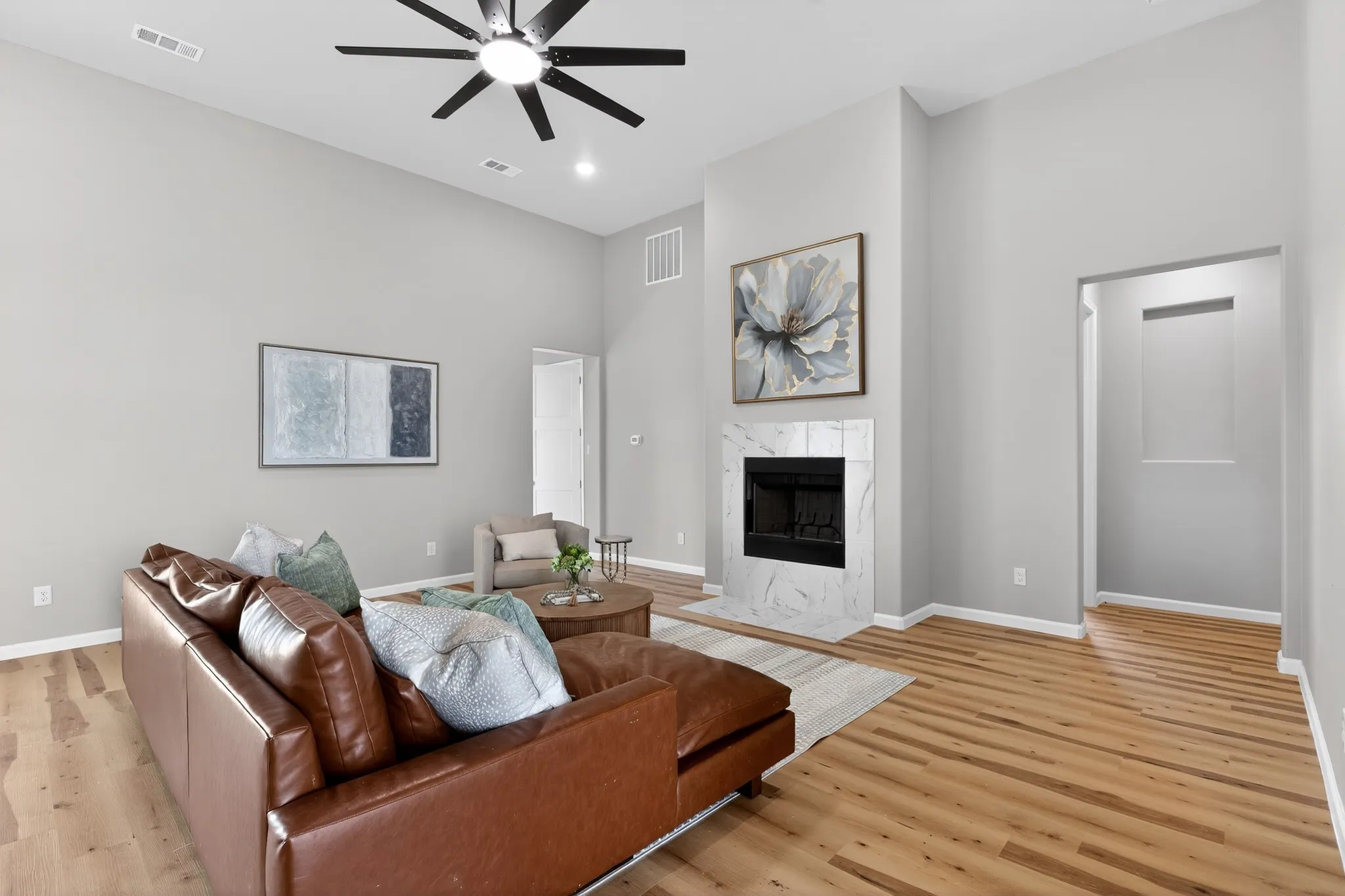 Spacious living area featuring high vaulted ceilings, light wood-finish flooring, a contemporary ceiling fan, and a fireplace with a white marble-patterned surround