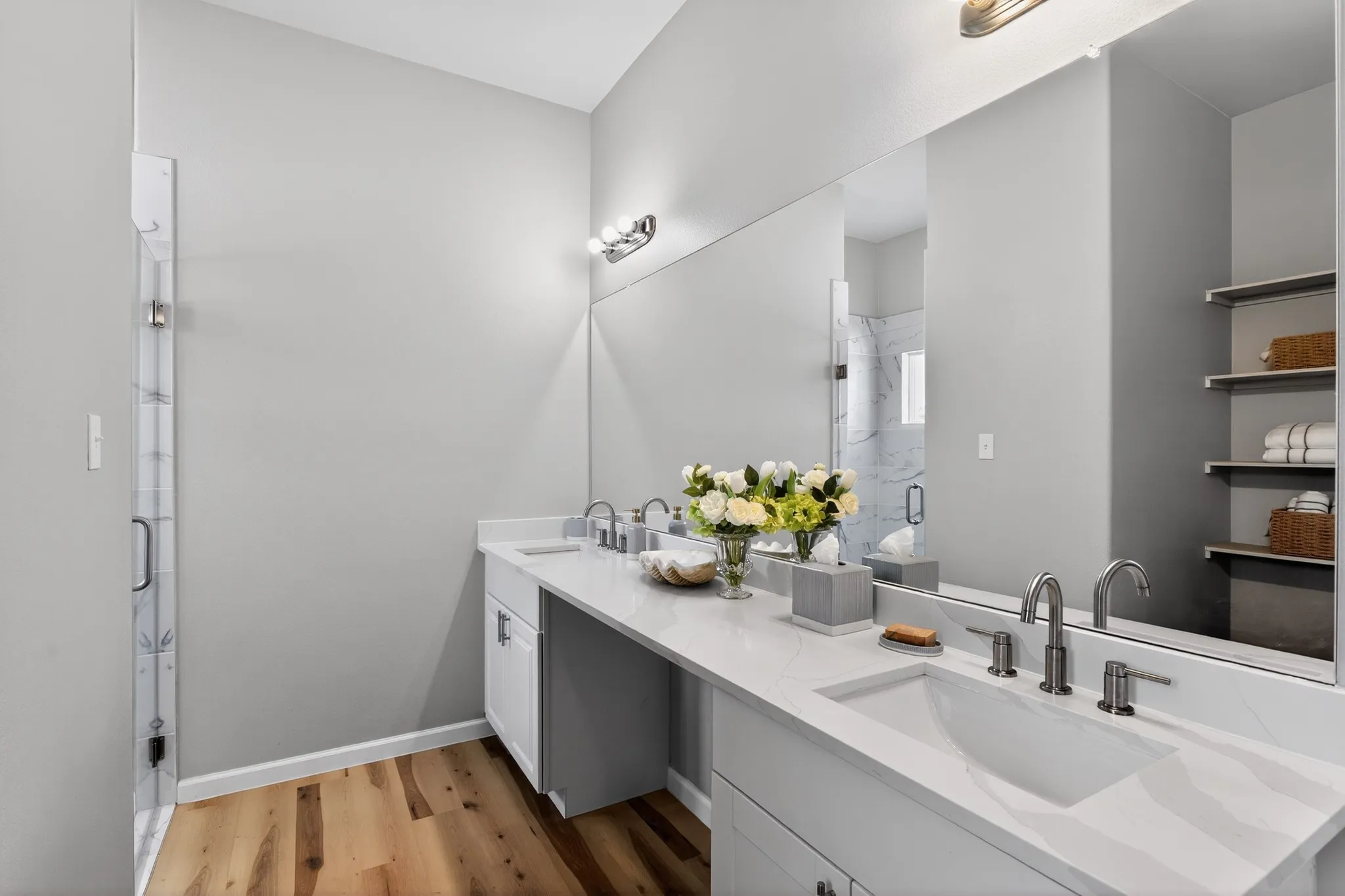 Spacious bathroom featuring a dual vanity with integrated sinks, light-toned countertops, wood-finish flooring, a full-width mirror, and a glass-enclosed shower with marble-look tile surround